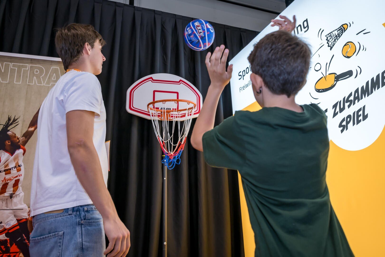 Zwei Jungen spielen drinnen an einem Messestand Basketball; einer wirft einen Ball auf einen kleinen Korb, während der andere in der Nähe steht. Im Hintergrund sind ein heller Bildschirm und ein Sportplakat zu sehen.