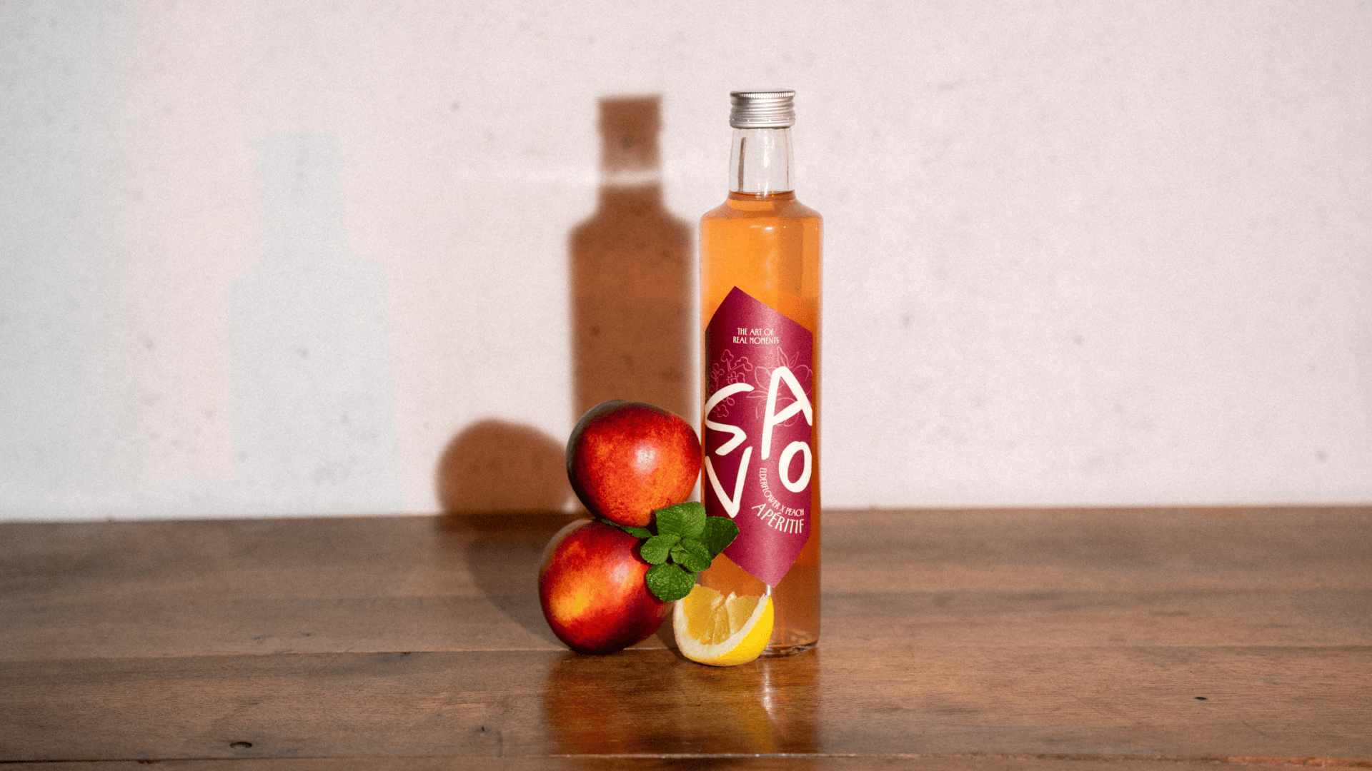 Eine Klarglasflasche mit orangefarbener Flüssigkeit und rotem Etikett steht auf einer Holzfläche. Davor befinden sich zwei rote Äpfel, eine Zitronenscheibe und ein Zweig Minze. Der Hintergrund ist einfarbig und leicht strukturiert.