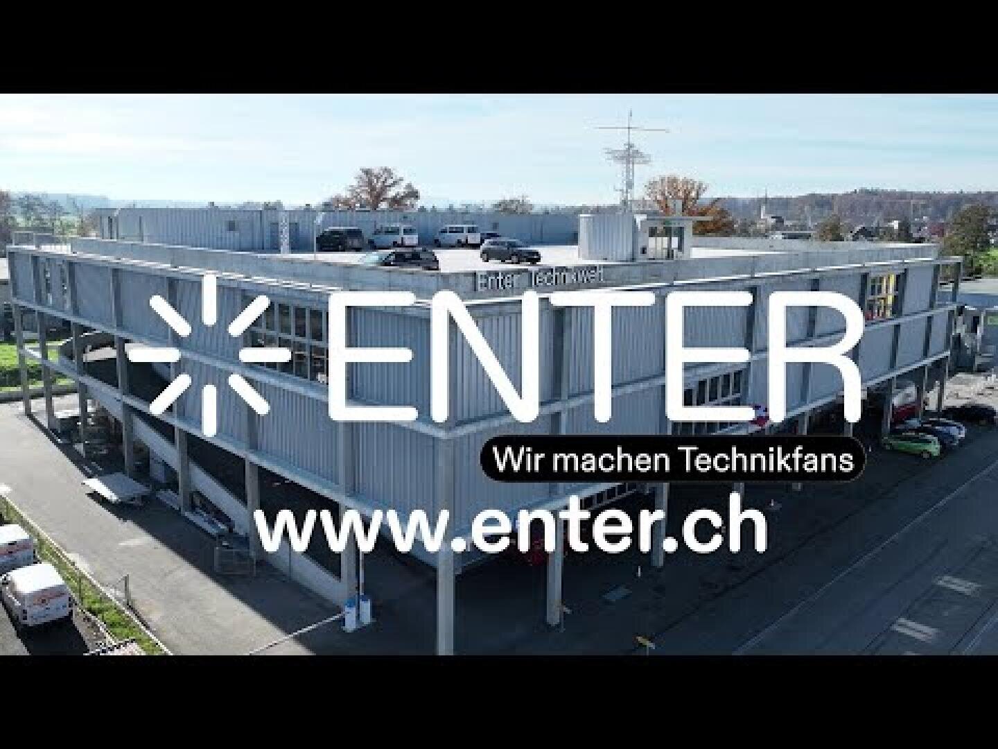 Ein großes, modernes Gebäude mit den Schriftzügen ENTER und Wir machen Technikfans sowie der Website www.enter.ch überlagert. Das Gebäude hat eine Dachfläche und ist von Bäumen und Autos umgeben.