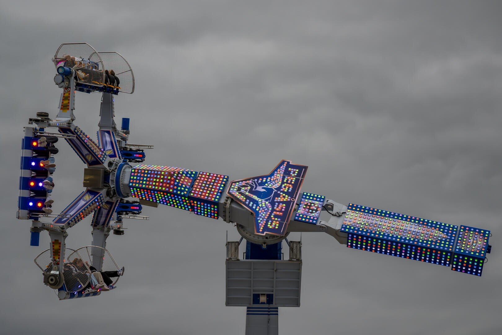 Ein hell beleuchtetes Fahrgeschäft in einem Vergnügungspark wirbelt Menschen in Sitzen hoch über dem Boden gegen einen bewölkten Himmel. Die Arme und die Basis des Fahrgeschäfts sind mit bunten LED-Leuchten ausgestattet.