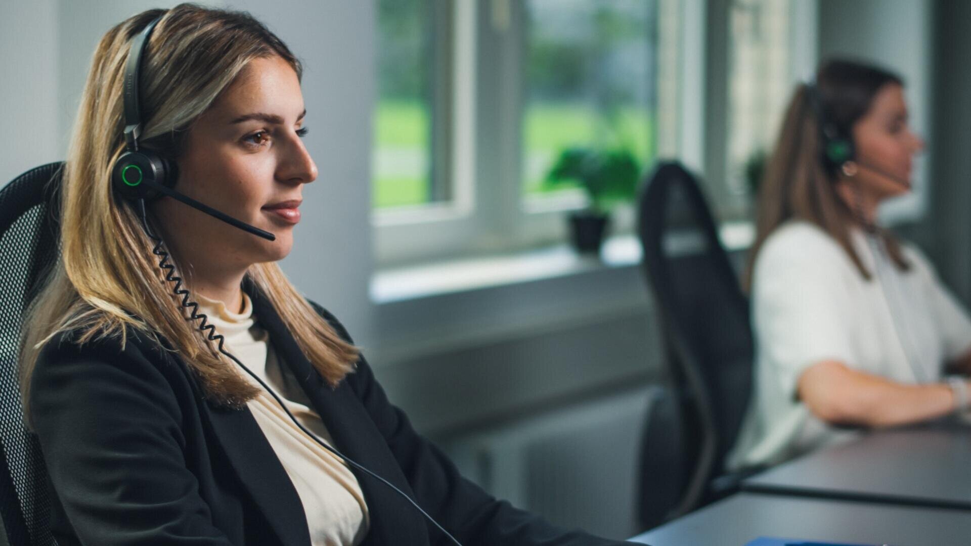 Zwei Frauen mit Headsets sitzen an Schreibtischen in einem Büro und arbeiten als Kundenbetreuerinnen. Der Hintergrund zeigt große Fenster mit natürlichem Licht und einem Blick ins Grüne.