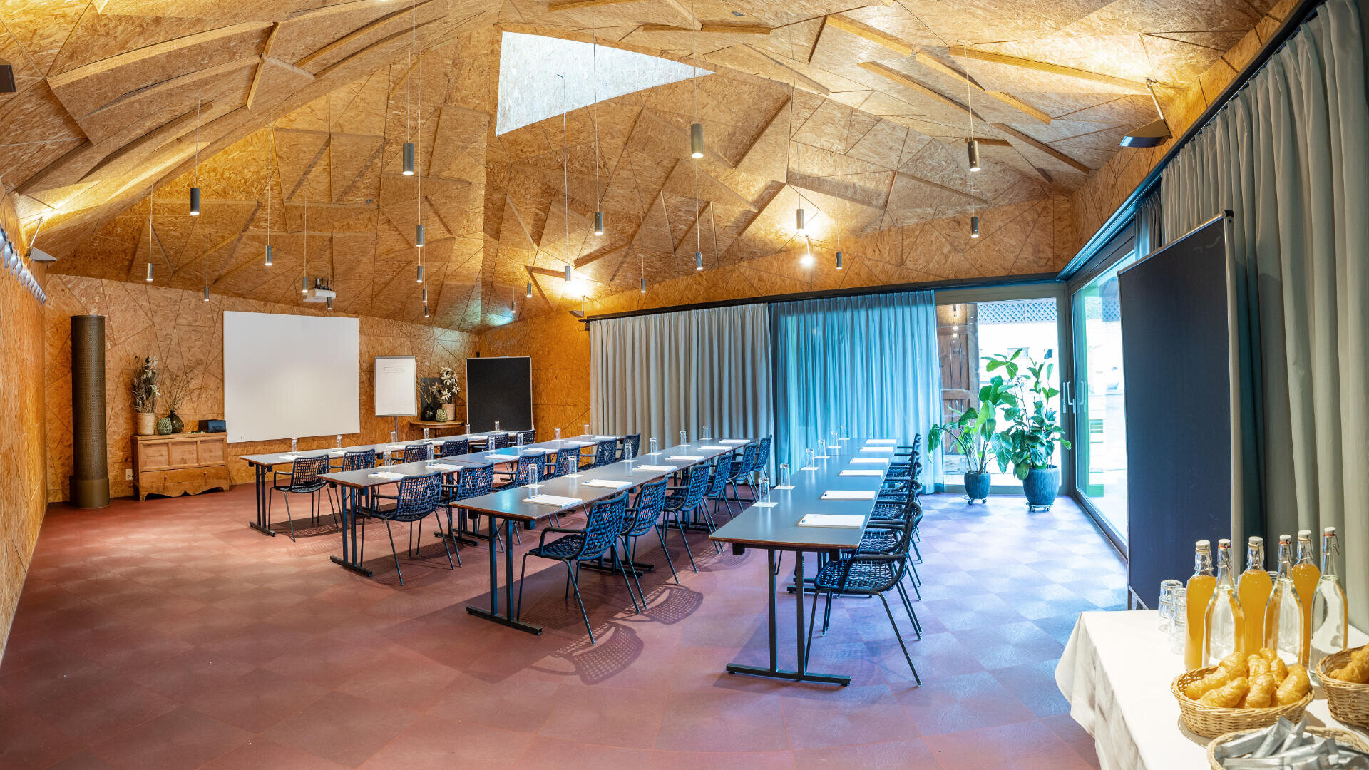 Ein moderner Konferenzraum mit einer geometrischen Holzdecke, Reihen von Tischen und Stühlen, einem Projektor, großen Fenstern mit Vorhängen, Topfpflanzen und einem Tisch mit Getränkeflaschen im Vordergrund.