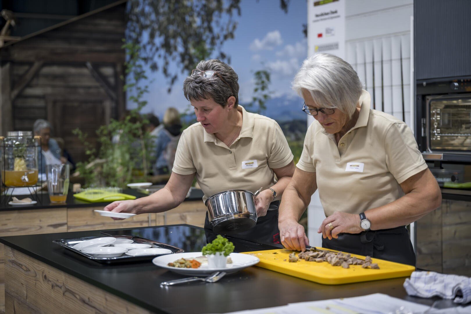 Zwei Frauen in beigen Hemden bereiten in einer modernen Küche Speisen zu. Eine schneidet Fleisch auf einem gelben Schneidebrett, während die andere Soße auf Teller gießt. Es gibt verschiedene Küchengeräte und einen unscharfen Hintergrund mit Grünzeug.