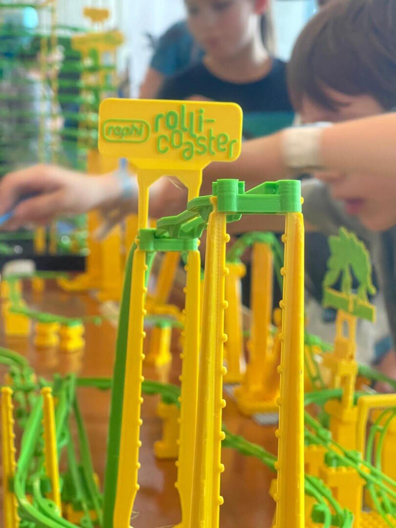 Nahaufnahme eines bunten Spielzeug-Achterbahn-Sets mit gelben und grünen Schienen, beschriftet mit rolli-coaster, während zwei Kinder im Hintergrund spielen und das Spielzeug zusammenbauen oder damit spielen.