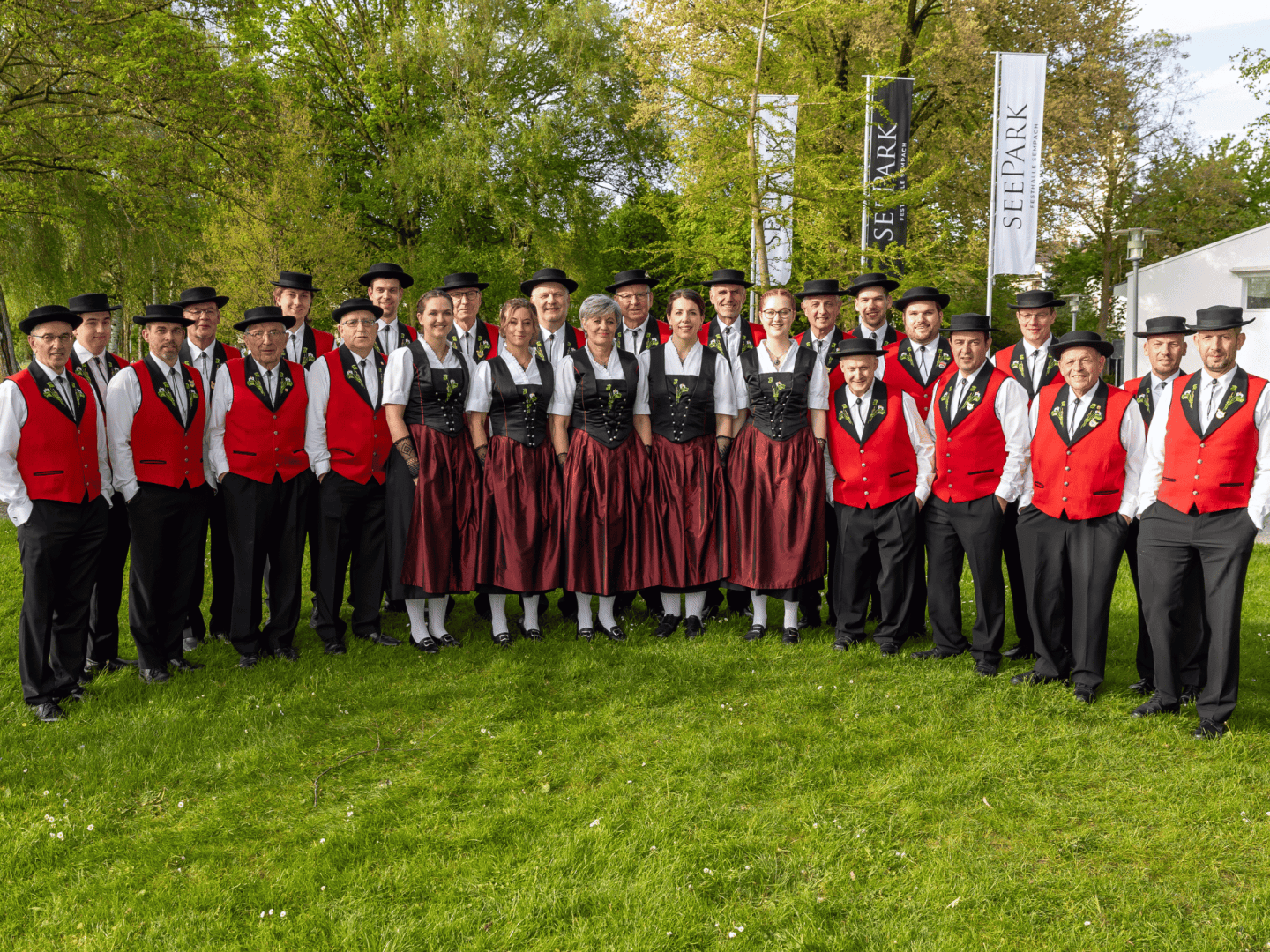 Eine Gruppe von Männern und Frauen in traditioneller roter und schwarzer Tracht posiert gemeinsam im Freien auf einer Wiese, mit Bäumen und zwei großen Bannern im Hintergrund.