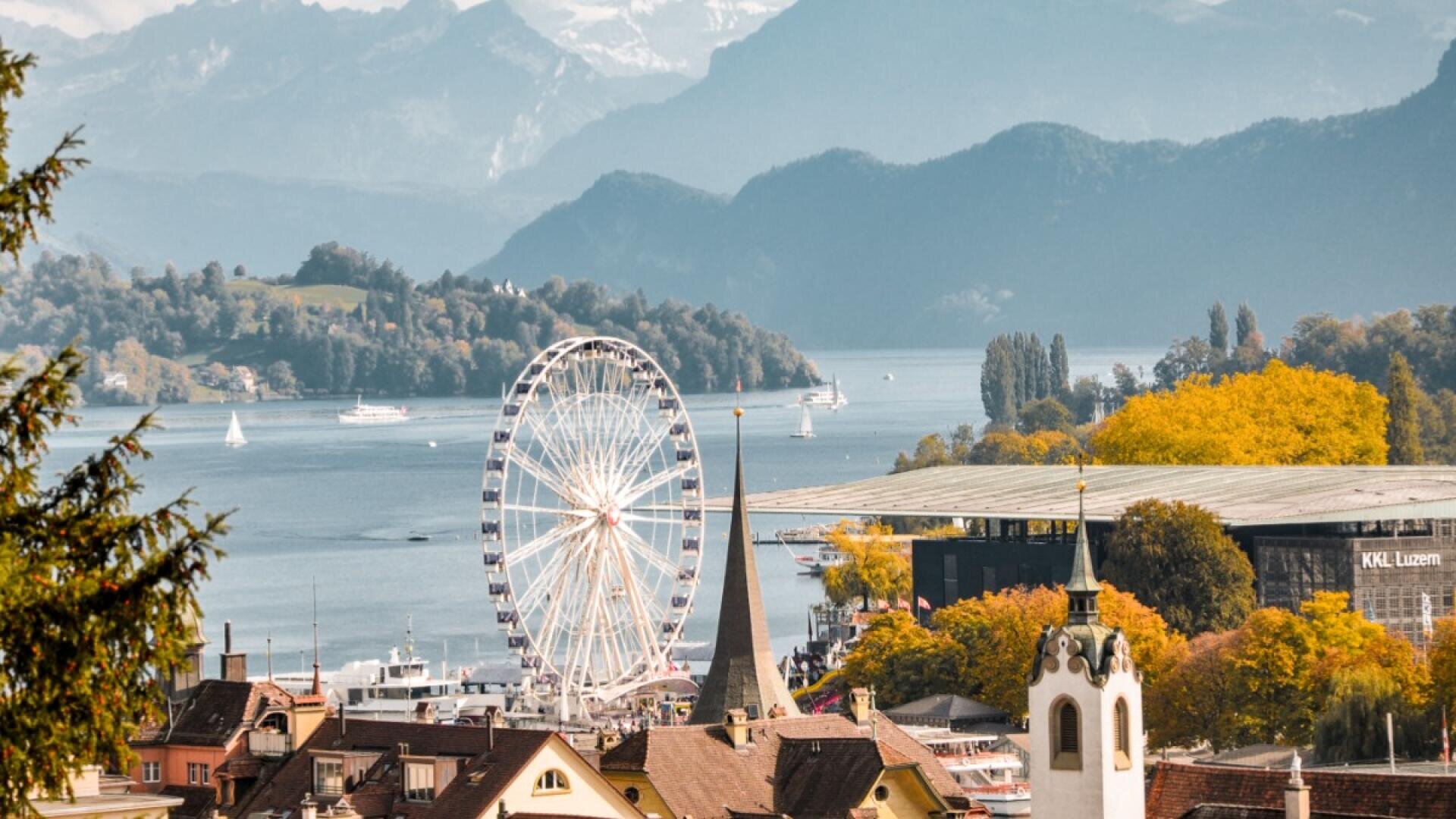 Eine malerische Stadt am See mit einem großen Riesenrad, einem Kirchturm und bunten Herbstbäumen vor der Kulisse von Bergen und einem ruhigen See mit Segelbooten.