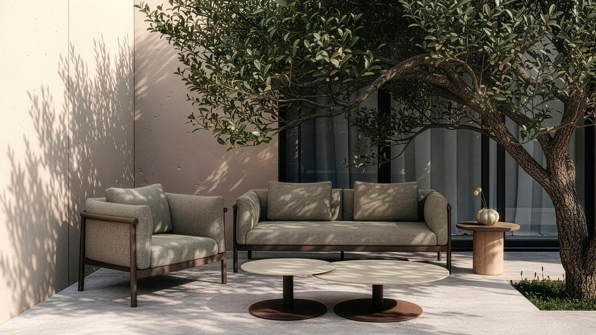 Moderne Außenterrasse mit einem gepolsterten Sessel, einem Sofa, zwei runden Couchtischen und einem kleinen Beistelltisch mit einer Vase, im Schatten eines Laubbaums neben einem Gebäude mit großen Fenstern.