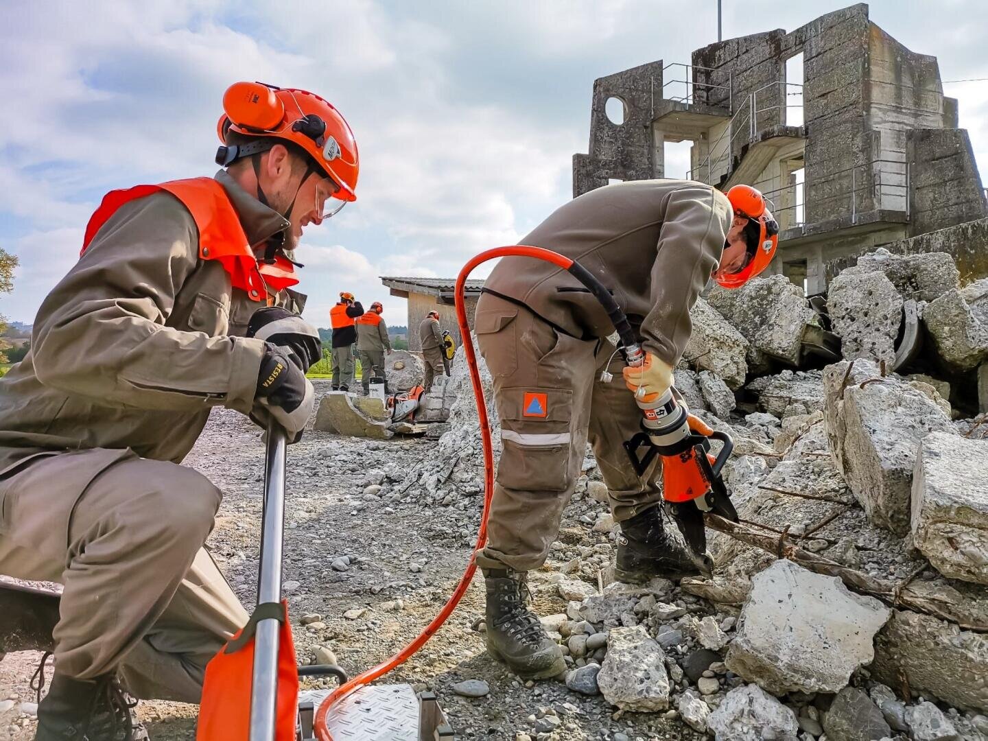 Zwei Rettungskräfte in orangefarbenen Helmen und Uniformen durchsuchen mit Werkzeugen Betontrümmer, während im Hintergrund weitere Teammitglieder und ein beschädigtes Gebäude zu sehen sind. Die Szene deutet auf einen Katastropheneinsatz oder eine Notfallübung hin.