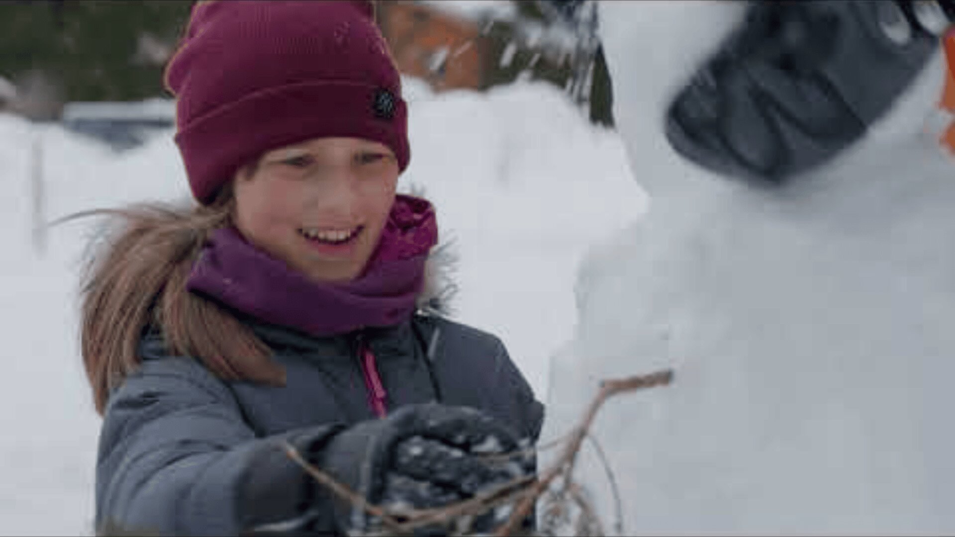 Ein lächelndes Kind mit kastanienbrauner Mütze und lilafarbenem Schal steht draußen im Schnee und streckt seine Hand aus, um beim Bau eines Schneemanns mit Stöcken als Arme zu helfen.