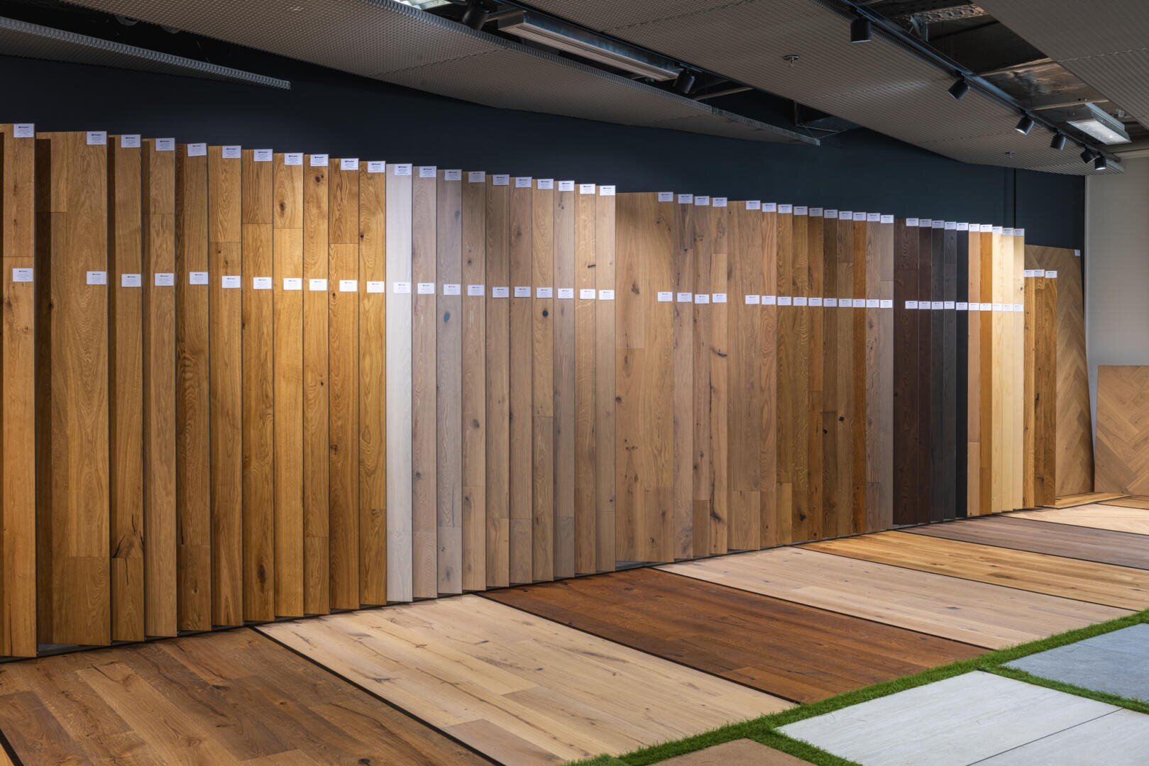 Ein Ausstellungsraum mit verschiedenen Holzfußbodenmustern, die vertikal an einer dunklen Wand angeordnet sind und verschiedene Holzfarben und Maserungen zeigen, mit zusätzlichen Bodenmustern, die horizontal darunter angeordnet sind.