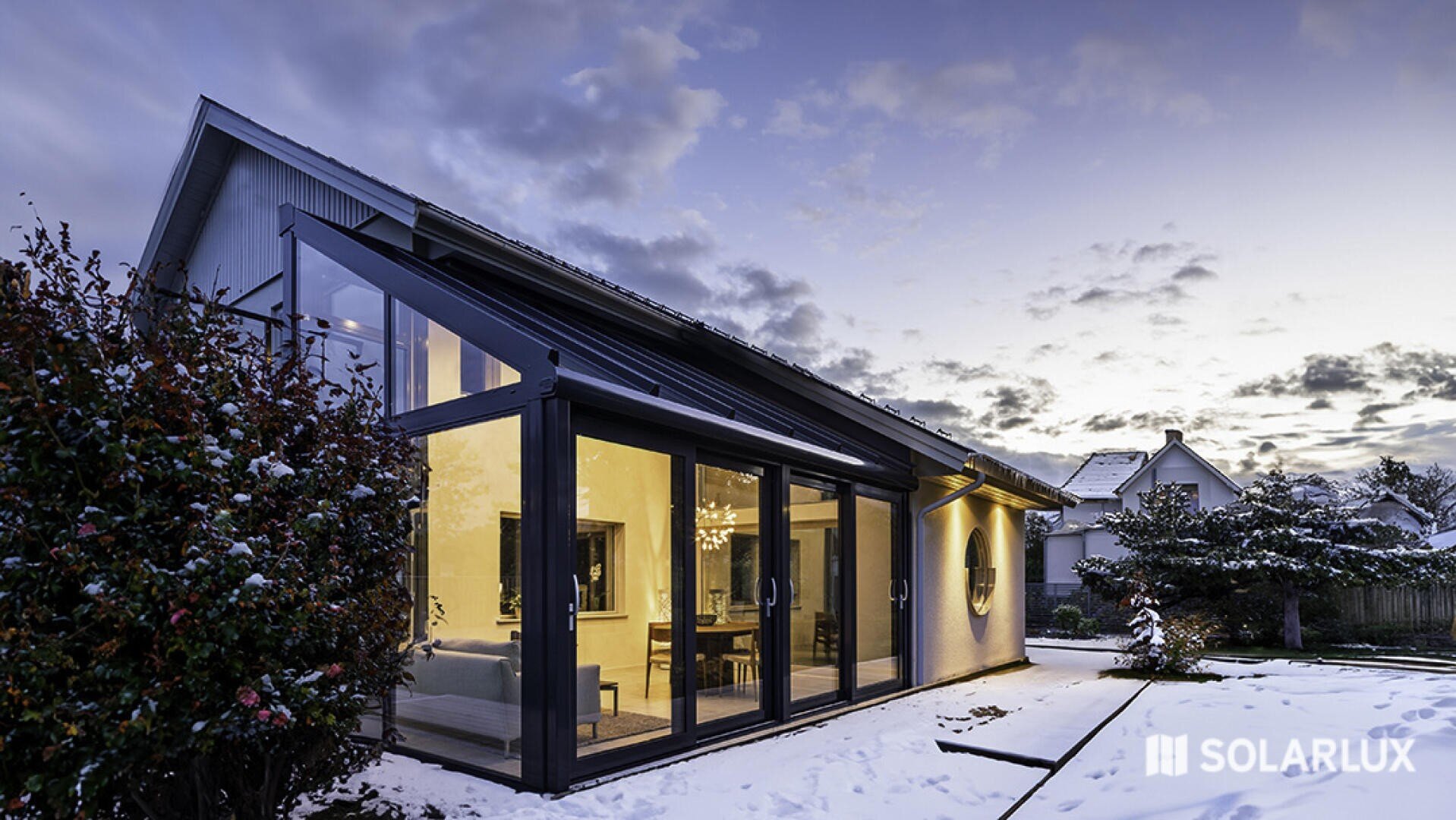 Modernes Haus mit großen Glasfenstern und einem schrägen Dach, von innen warm beleuchtet. Schnee bedeckt den Boden und die Büsche, und der Himmel ist bei Sonnenuntergang teilweise bewölkt. Das Solarlux-Logo ist in der unteren rechten Ecke zu sehen.
