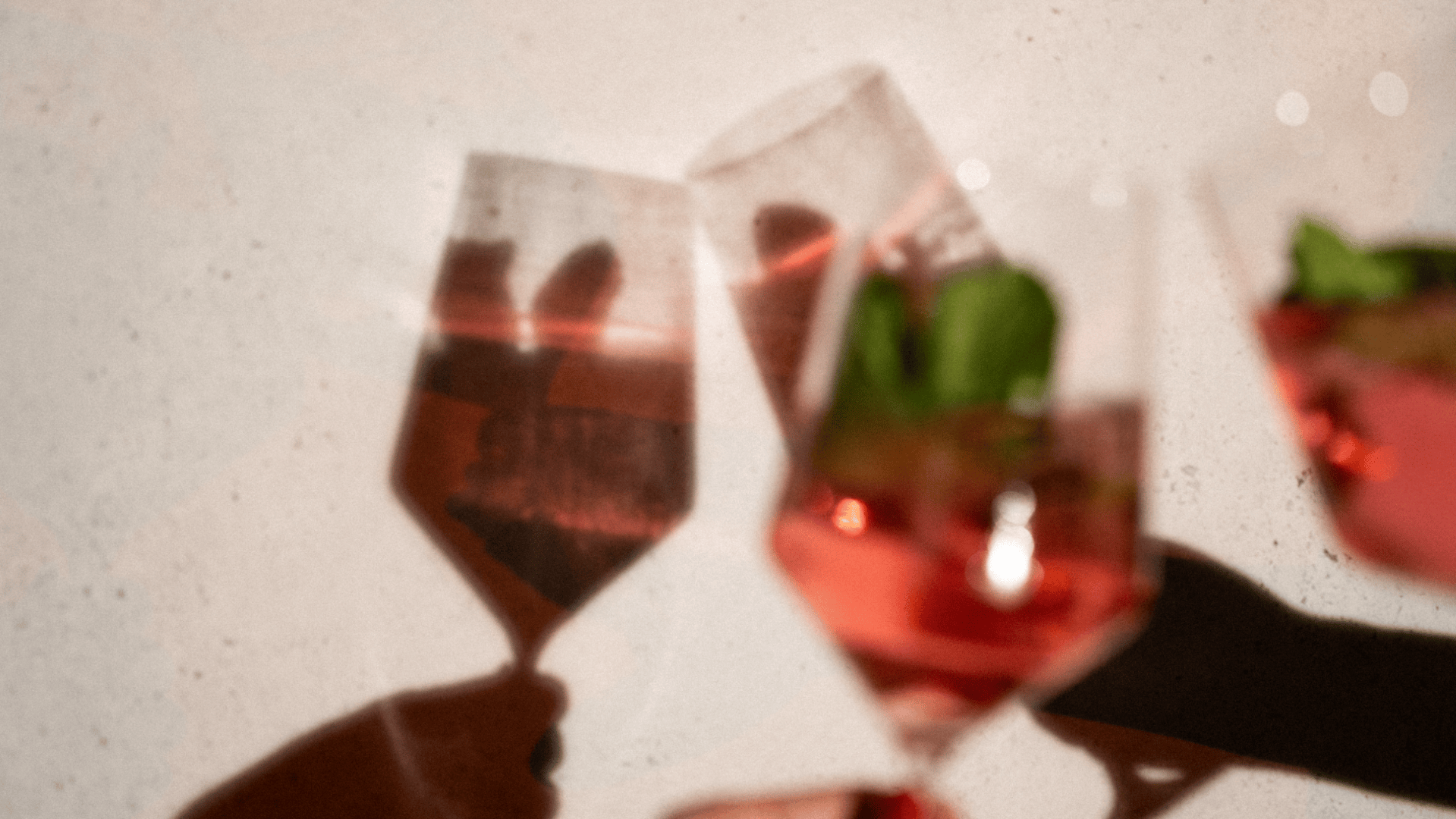 Drei mit einem rosafarbenen Getränk gefüllte und mit grünen Kräutern garnierte Gläser werden aneinandergehalten und werfen weiche Schatten auf eine helle Wand. Das Bild hat eine warme, feierliche Atmosphäre.