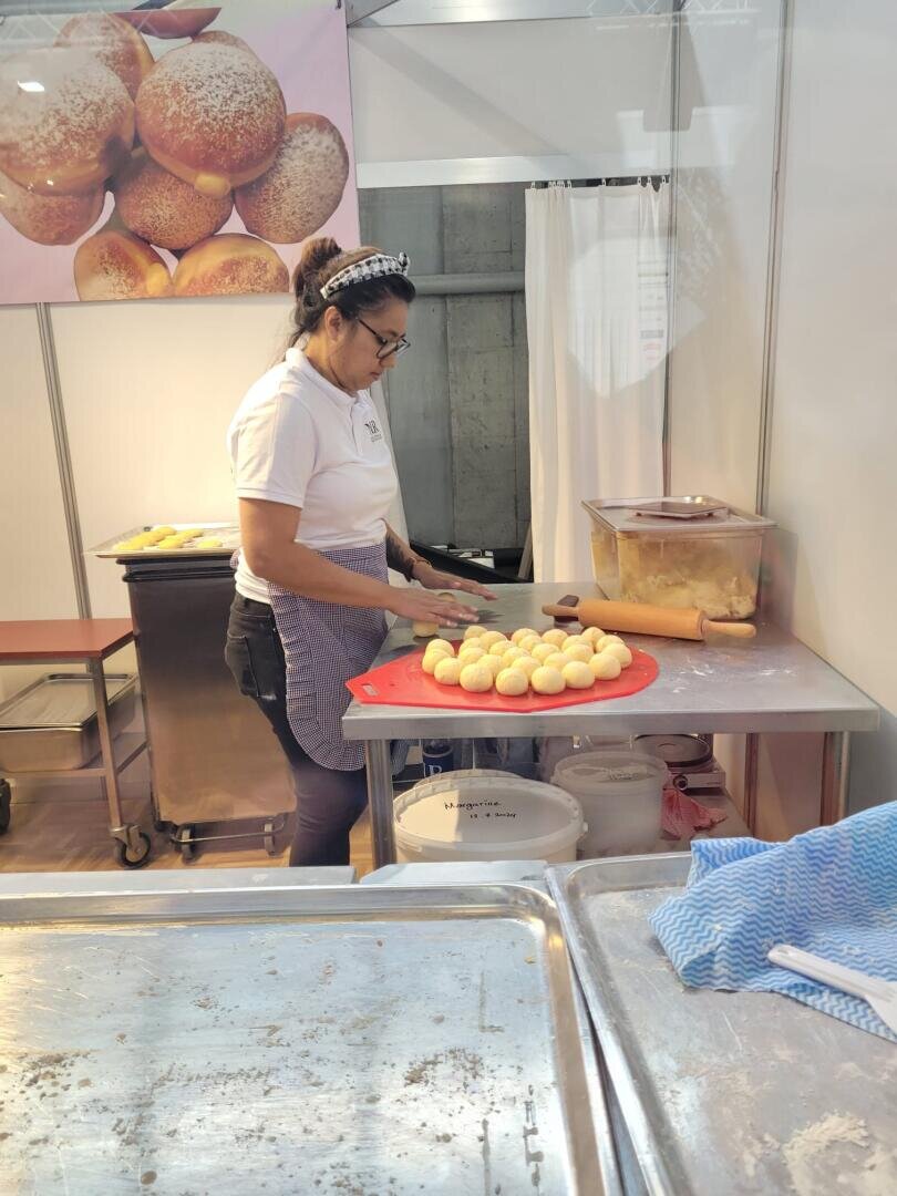 Eine Frau mit Brille und Schürze formt Teigkugeln auf einem roten Schneidebrett in einem Bäckereistand. Hinter ihr steht ein Poster mit runden Broten und Behältern mit Zutaten auf der Theke.