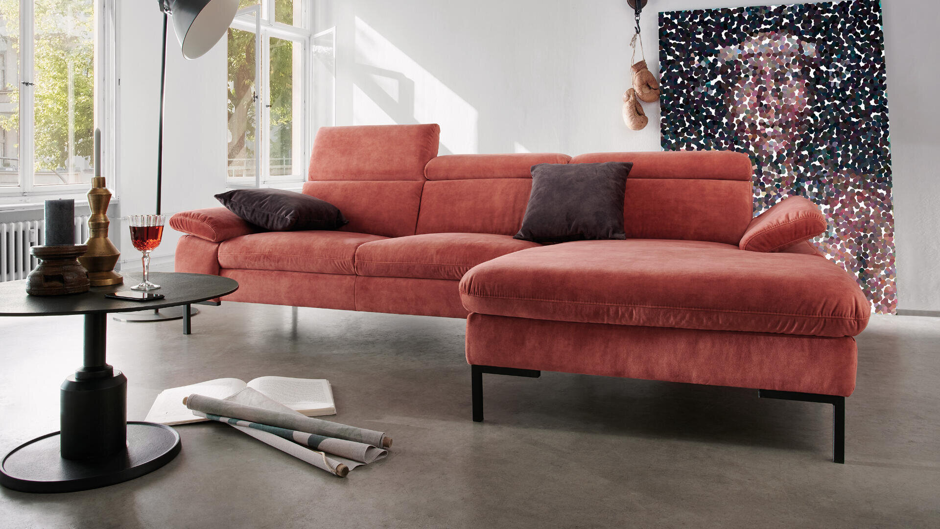 Modernes Wohnzimmer mit einem rosa Ecksofa, schwarzen Beinen, schwarzen und violetten Kissen, einem runden Couchtisch mit Deko und einem Glas mit einem roten Getränk, Büchern und Papieren auf dem Boden sowie abstrakter Kunst an der weißen Wand.