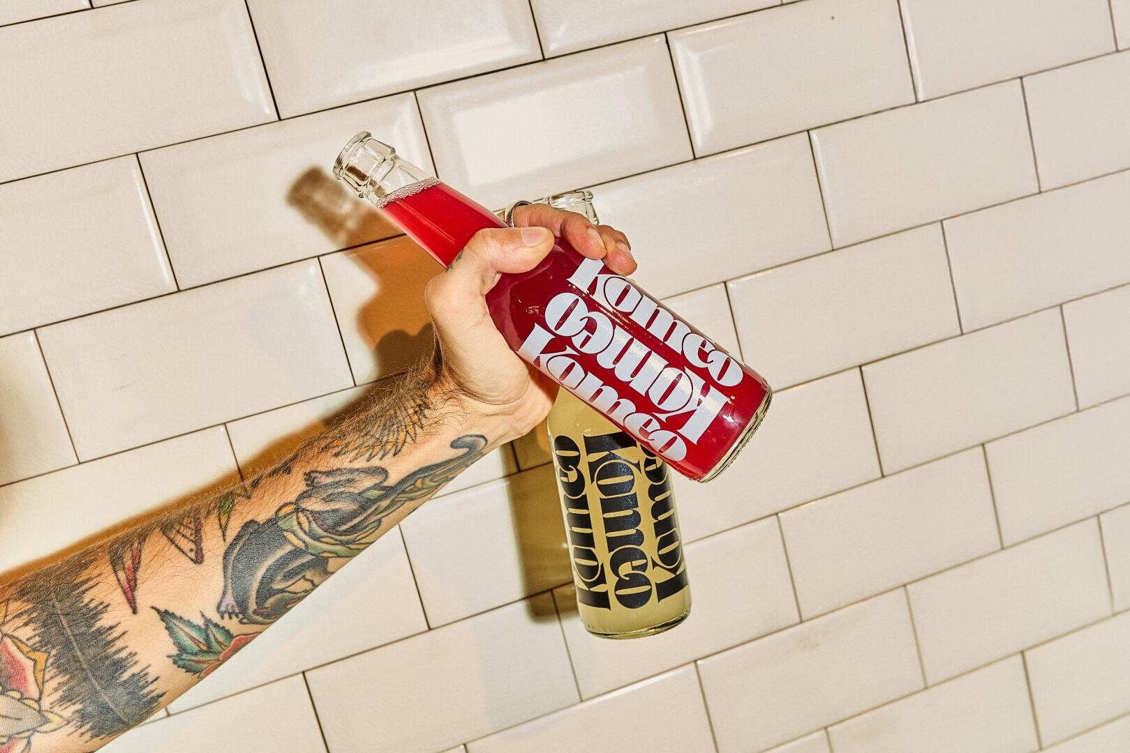 Ein tätowierter Arm hält vor einem gekachelten Wandhintergrund zwei Flaschen mit fettgedruckten, stilisierten Etiketten, von denen eine mit einer roten und die andere mit einer klaren gelben Flüssigkeit gefüllt ist.