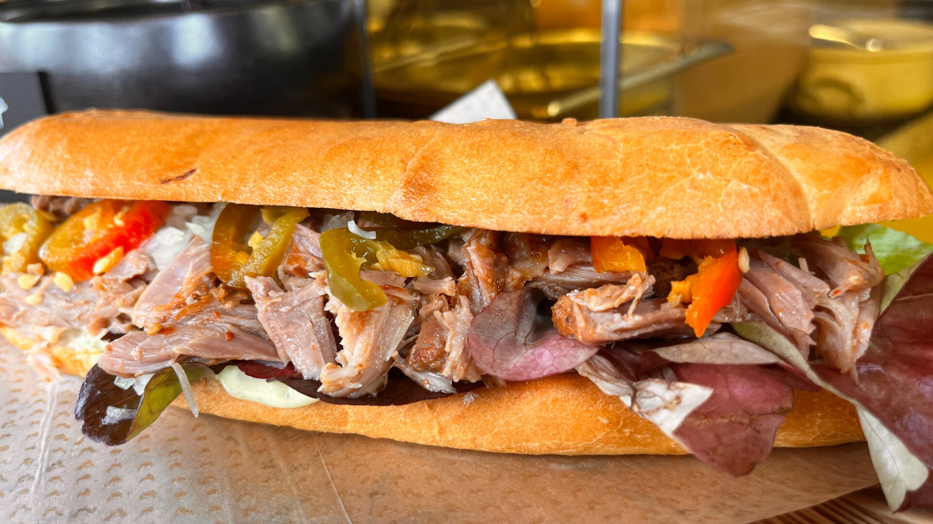 Nahaufnahme eines Baguette-Sandwichs, gefüllt mit geschnetzeltem Fleisch, gemischtem Blattsalat, Jalapeño-Scheiben und roten Paprikaschoten, auf einem Stück braunem Backpapier.