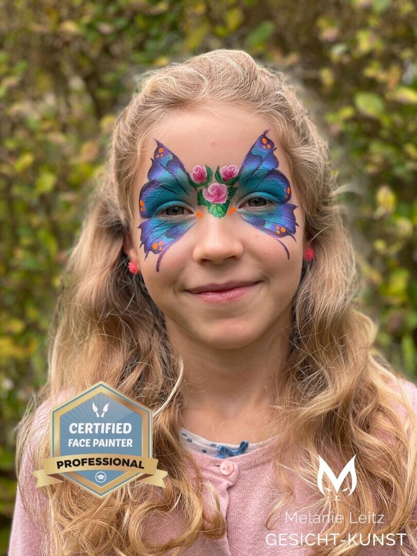 Ein junges Mädchen mit langen blonden Haaren lächelt, ihr Gesicht ist mit einem bunten Schmetterling und Blumen bemalt. Sie steht im Freien mit Grünpflanzen im Hintergrund. Ein Certified Face Painter Professional Abzeichen und ein Logo sind über das Bild gelegt.