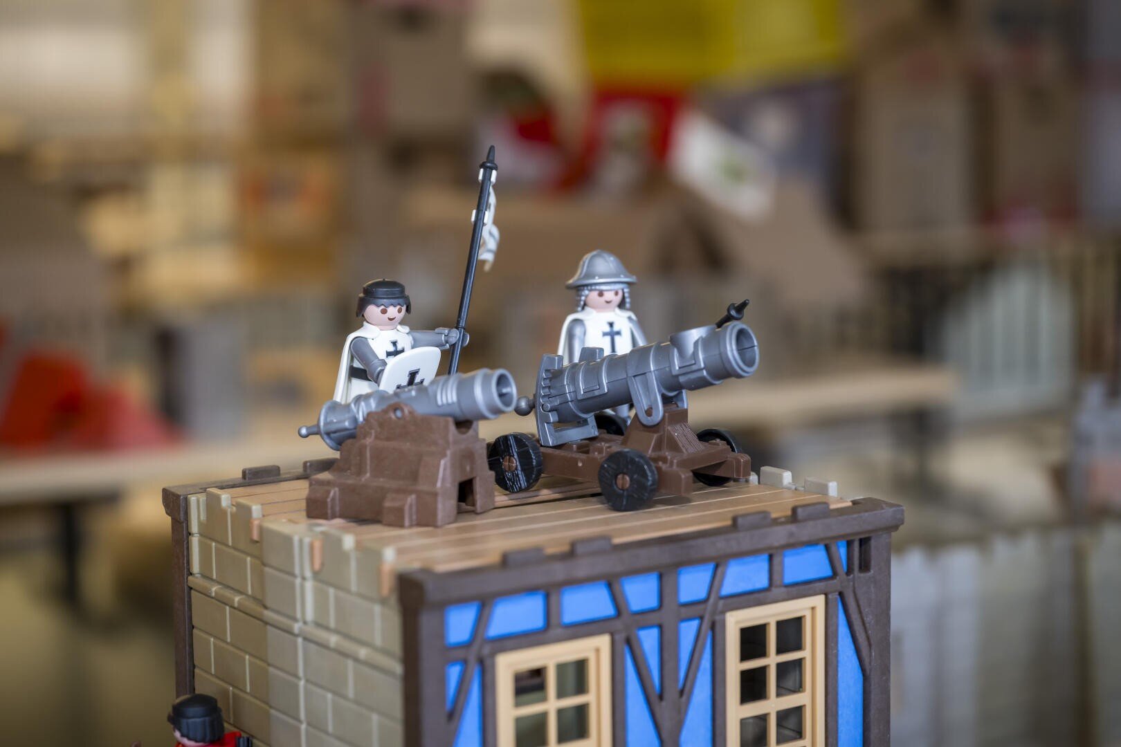 Zwei Playmobil-Ritter in Rüstung, einer mit einer Fahne in der Hand, stehen hinter Kanonen auf einem kleinen Modellgebäude mit blauen Akzenten, das einer mittelalterlichen Festung ähnelt. Der Hintergrund ist unscharf.