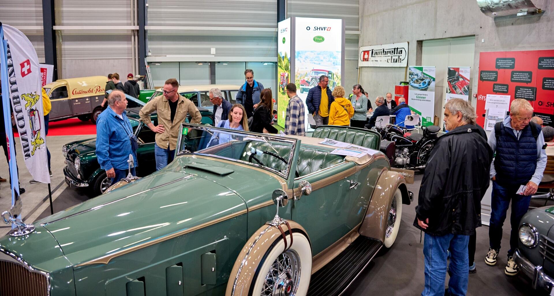 Menschen versammeln sich um ein grünes Oldtimer-Cabriolet, das auf einer Autoausstellung ausgestellt ist, während im Hintergrund Informationsschilder, Banner und andere klassische Fahrzeuge zu sehen sind.