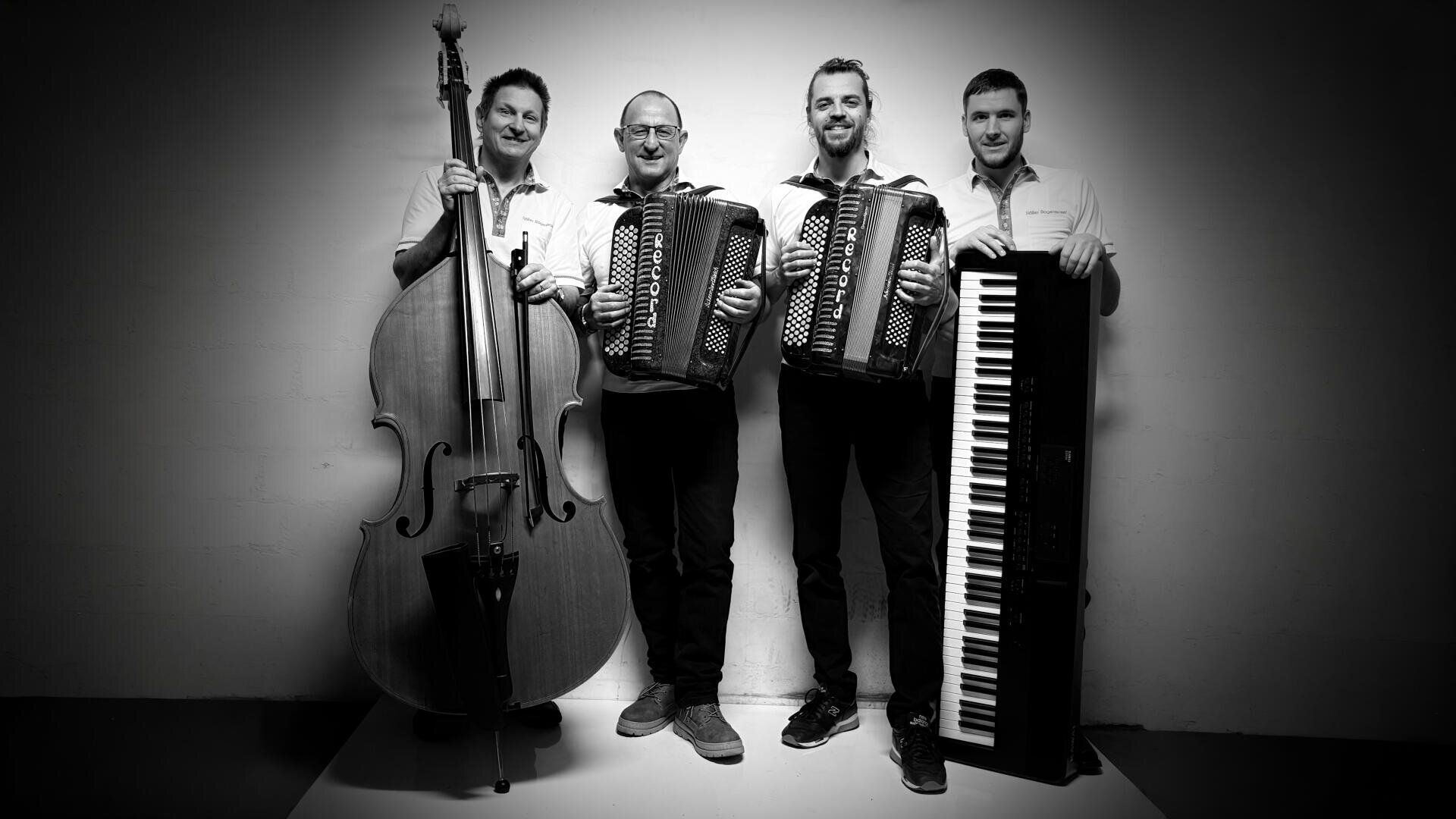 Vier Männer stehen lächelnd nebeneinander und halten Musikinstrumente, darunter einen Kontrabass, zwei Akkordeons und ein Keyboard, vor einem schlichten Hintergrund mit Scheinwerfereffekt.