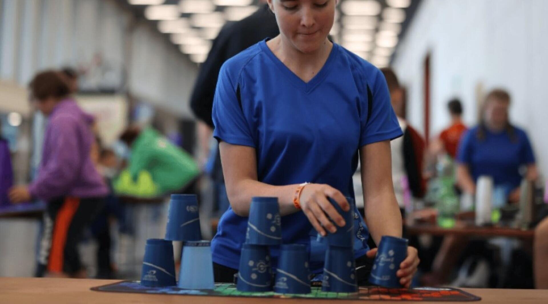 Eine Person in einem blauen Hemd stapelt schnell blaue Plastikbecher auf einem Tisch, bei dem es sich um einen Becherstapelwettbewerb zu handeln scheint, wobei andere Teilnehmer im Hintergrund zu sehen sind.