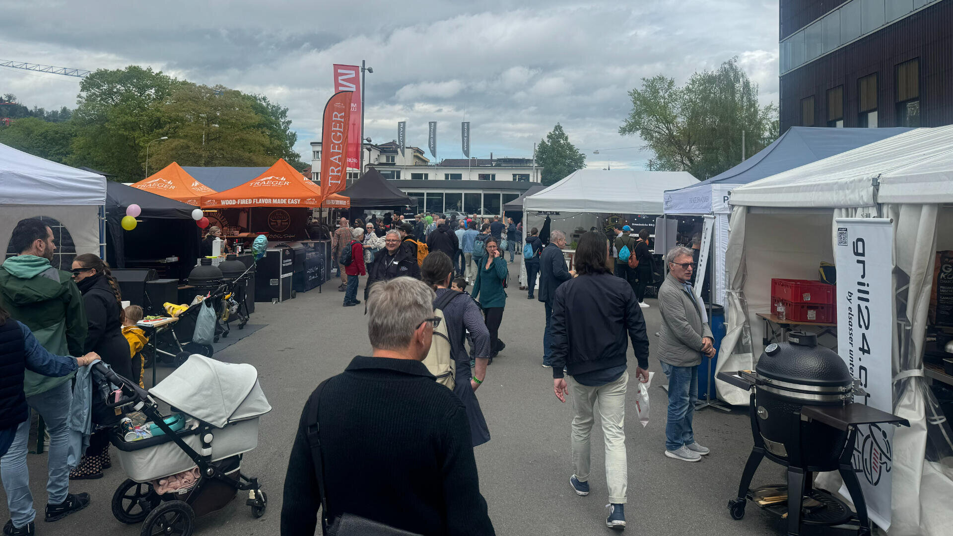 An einem bewölkten Tag spazieren die Menschen über einen Markt mit weißen und orangefarbenen Verkaufszelten. Einige schieben Kinderwagen, während andere die Stände durchstöbern. Im Hintergrund sind Gebäude und Bäume zu sehen.