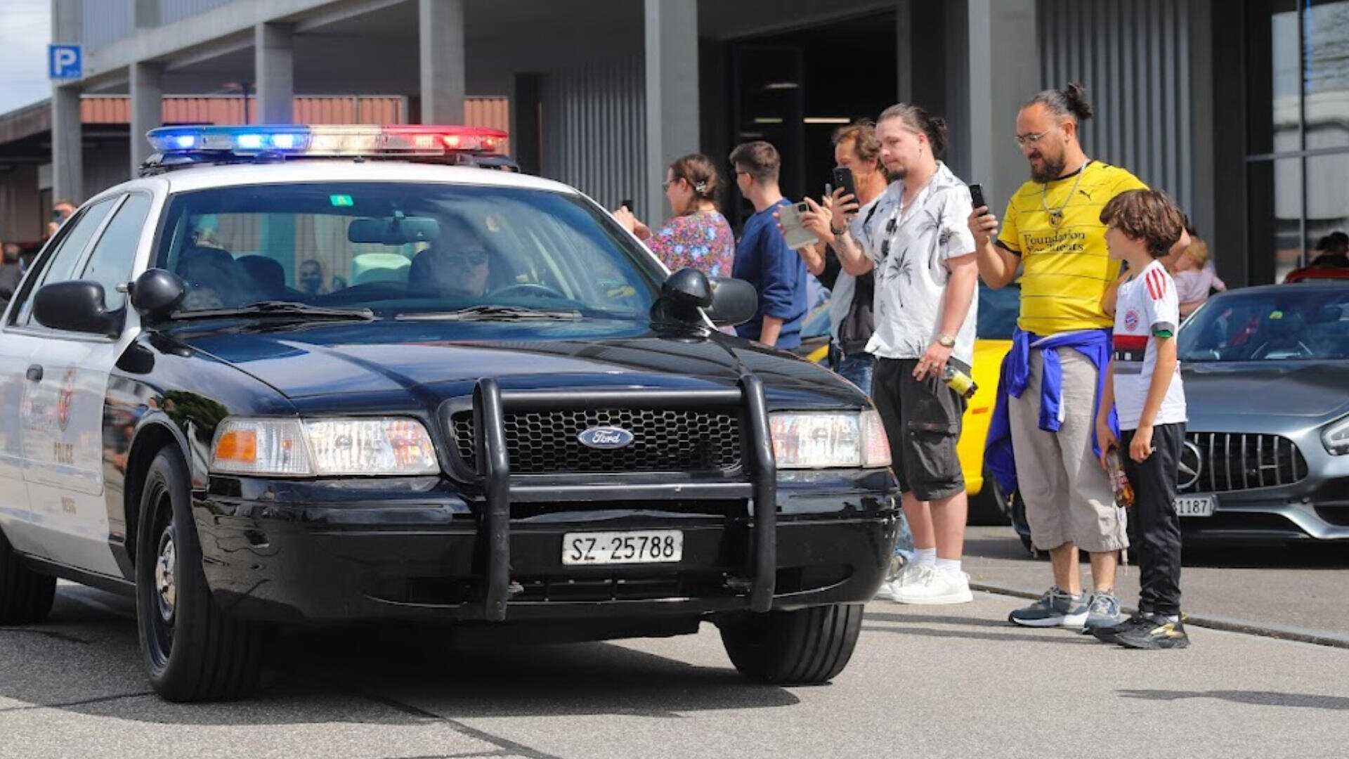 Ein schwarzes Polizeiauto mit Blinklichtern parkt auf einer Straße, während mehrere Personen in der Nähe Fotos machen und zusehen; ein Mann und ein Kind stehen am nächsten, im Hintergrund sind Sportwagen und ein Gebäude zu sehen.