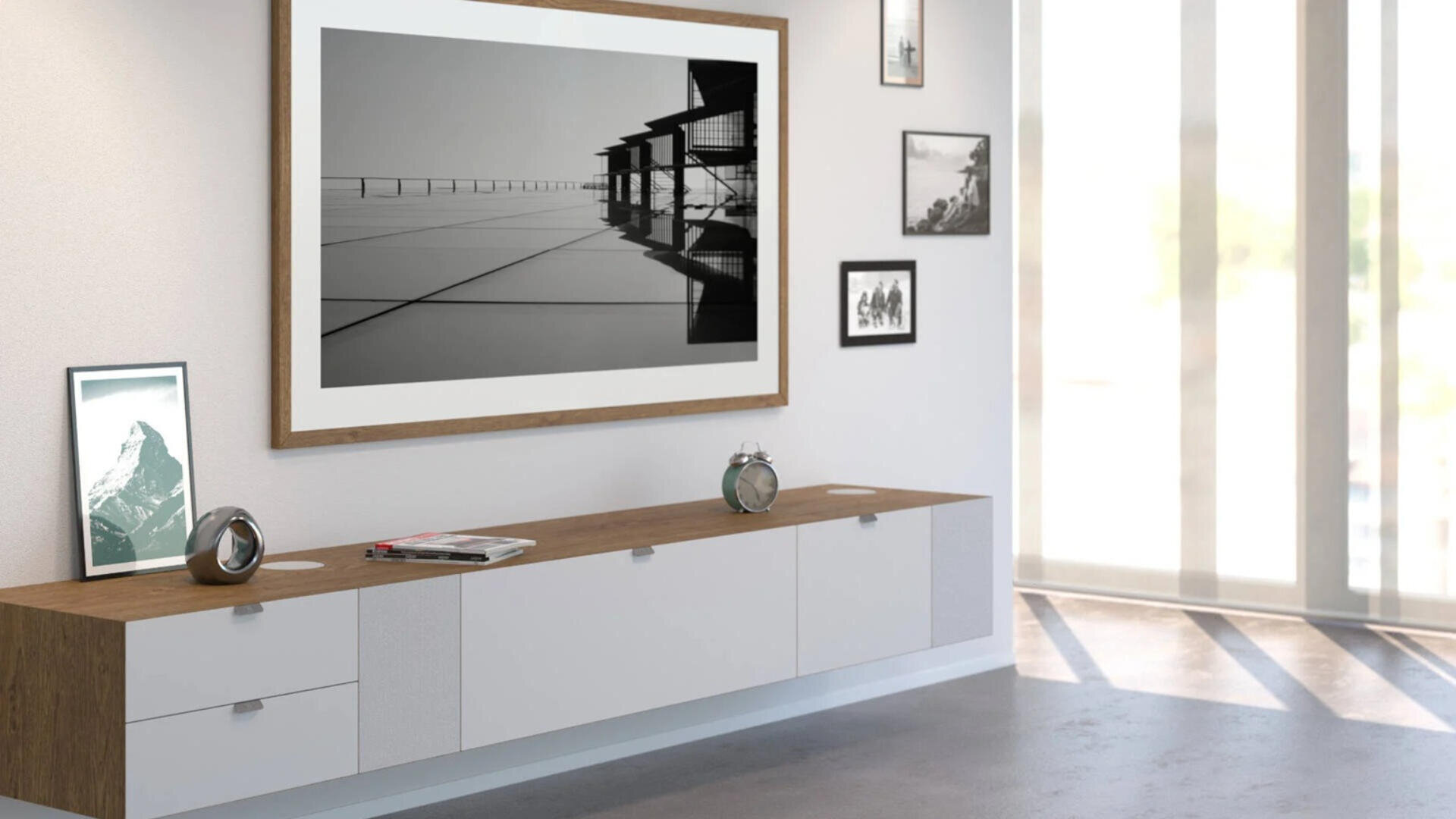 Ein modernes Wohnzimmer mit einem langen weißen und hölzernen Schrank, gerahmten Fotos und Kunstwerken an der Wand und Sonnenlicht, das durch große Fenster auf einen Betonboden fällt.