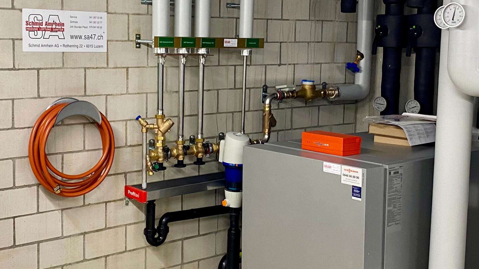 Eine Heizungsanlage mit Rohren, Ventilen, Messgeräten und einem Metallgerät vor einer gekachelten Wand. Ein orangefarbener Schlauch ist an der Wand aufgerollt, und in der oberen linken Ecke ist ein Schild mit Kontaktinformationen zu sehen.