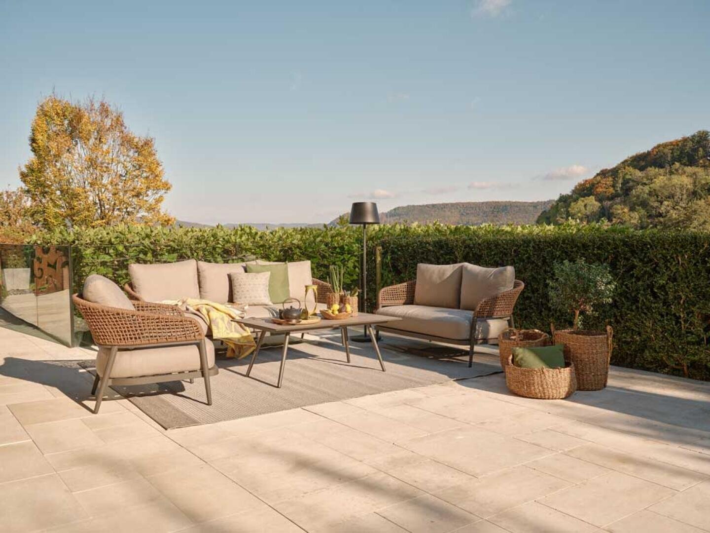 Eine moderne Außenterrasse mit gepolsterten Sofas, einem Couchtisch, einem Teppich, einer Stehlampe und geflochtenen Körben, umgeben von grünen Hecken und mit Blick auf ferne Hügel unter einem klaren blauen Himmel.