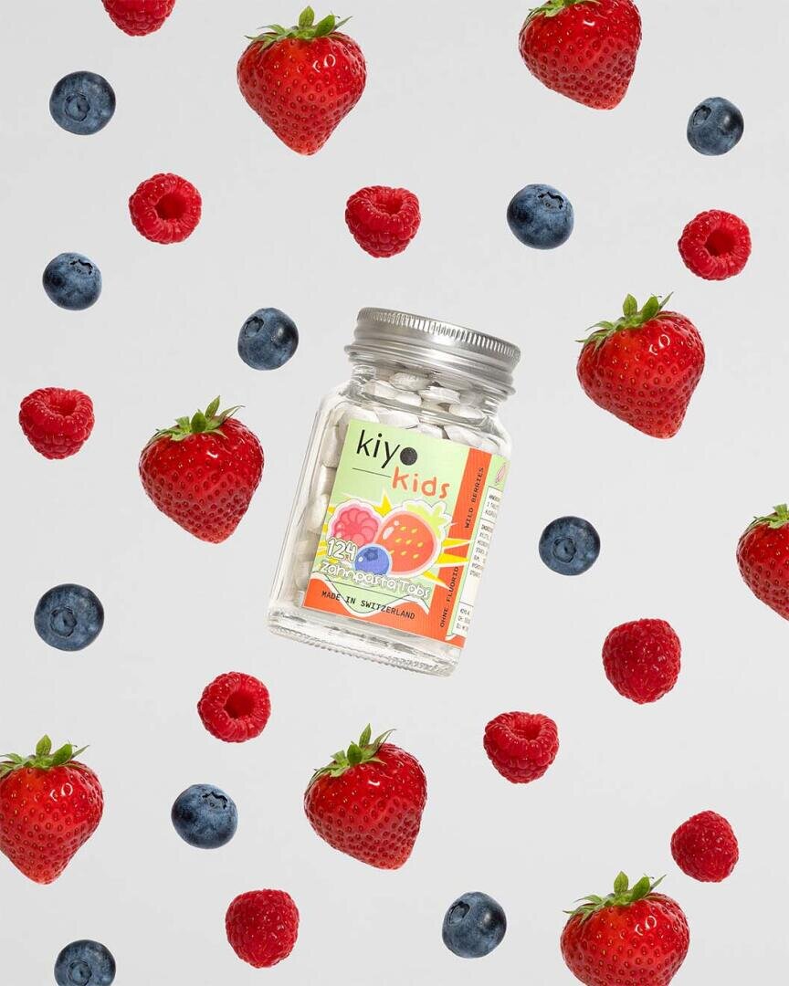 Ein Glas mit Kiyo Kids Vitaminen ist umgeben von schwimmenden Erdbeeren, Himbeeren und Blaubeeren auf weißem Hintergrund. Die farbenfrohen Früchte und die Flasche bilden eine verspielte, lebendige Szene.
