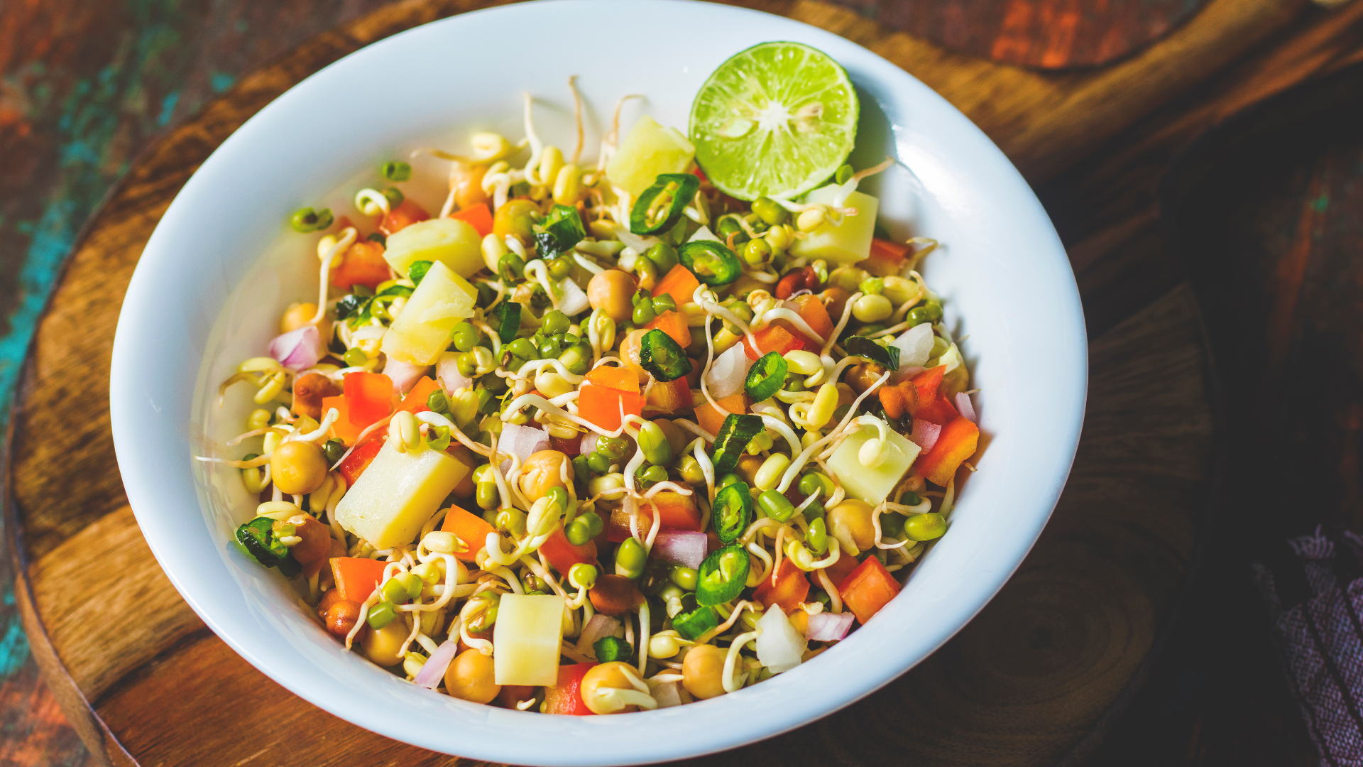 Eine weiße Schale, gefüllt mit einem bunten Sprossensalat, mit gehacktem Gemüse wie Tomaten, Zwiebeln, Gurken, Kartoffeln, grünen Chilis und einer halbierten Limette, die auf einem Holzbrett platziert wird.