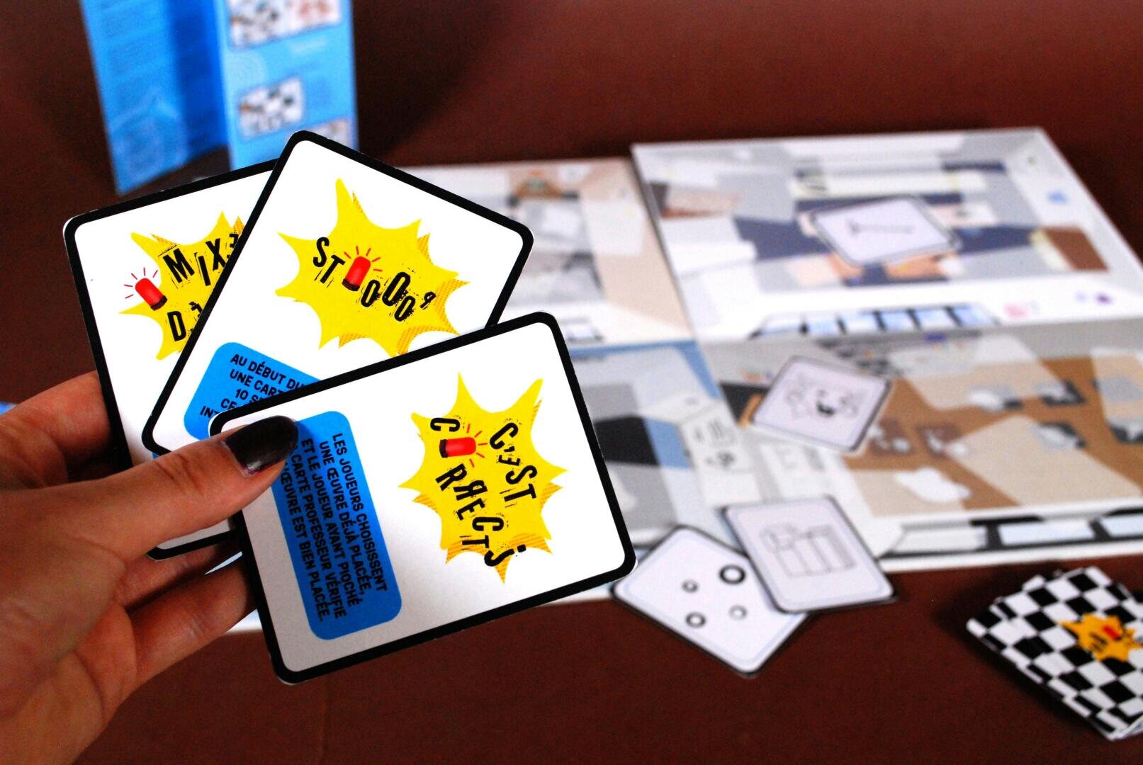 Eine Hand hält drei Spielkarten mit buntem Text im Comic-Stil in französischer Sprache über einem Brettspiel mit verschiedenen Karten und Spielsteinen im Hintergrund.
