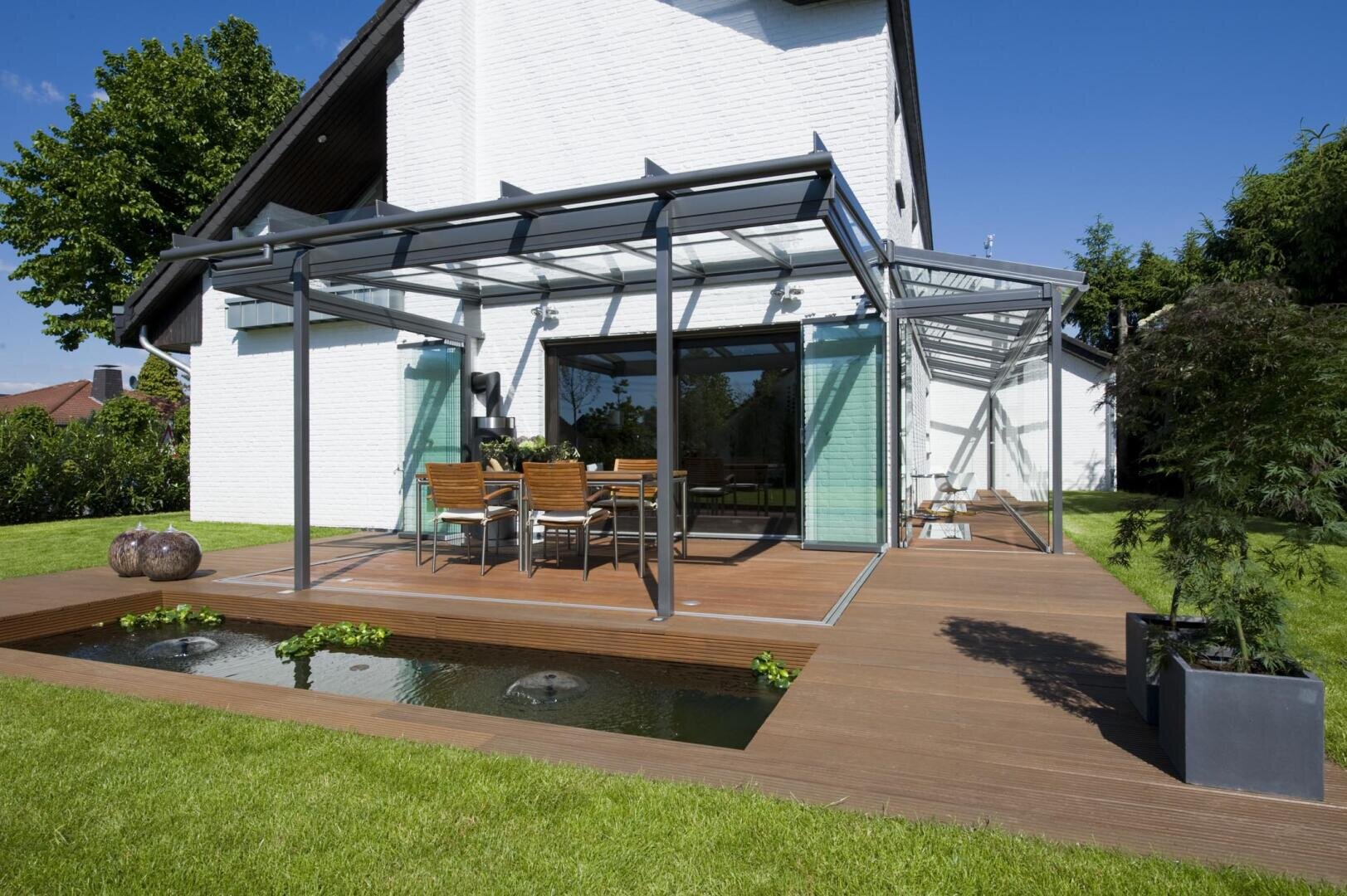 Eine moderne gläserne Terrassenüberdachung mit einem Esstisch und Stühlen befindet sich auf einer Holzterrasse neben einem flachen Teich, der an ein weißes Haus angebaut ist, umgeben von grünem Gras und Bäumen unter einem klaren blauen Himmel.