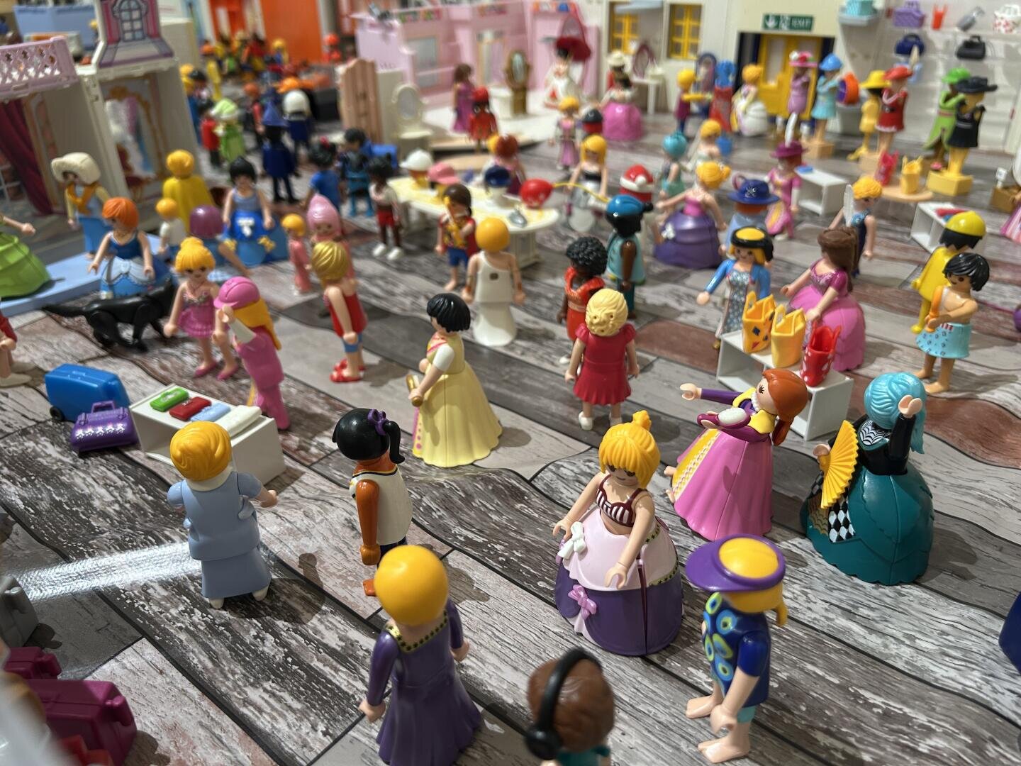 Bunte Spielzeugfiguren, meist Frauen und Kinder in Kleidern, sind in einer belebten Innenraumszene mit Möbeln und Auslagen versammelt, die den Eindruck eines lebhaften Marktes oder einer Veranstaltung erweckt. Der Boden besteht aus einem Holzmuster, und der Hintergrund ist detailliert.