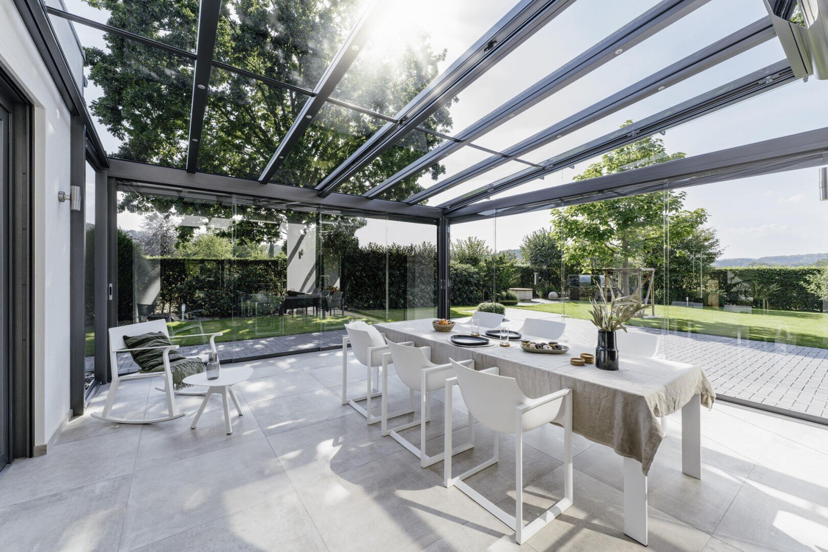 Eine moderne, verglaste Terrasse mit einem Esstisch, sechs weißen Stühlen und einer gemütlichen Sitzecke, die an einem sonnigen Tag einen Blick auf einen grünen Garten mit Bäumen und einer Rasenfläche bietet.