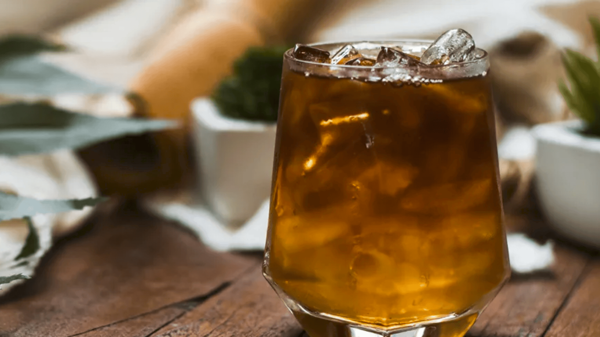 Ein Glas Eiskaffee oder Tee steht auf einem Holztisch, gefüllt mit Eiswürfeln. Der Hintergrund ist mit Topfpflanzen und einer gerollten Serviette sanft verschwommen.