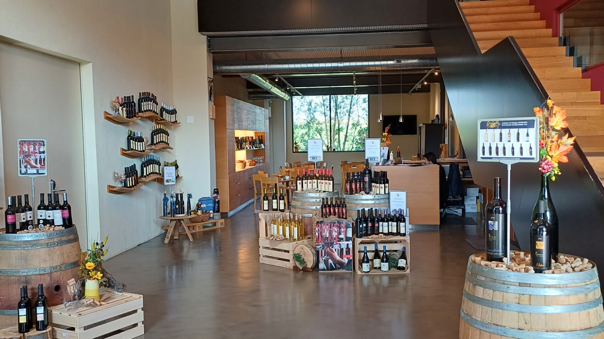 Eine moderne Weinhandlung mit Weinflaschen, die in Holzregalen, Fässern und Kisten ausgestellt sind. Es gibt Schilder mit Informationen, eine Treppe auf der rechten Seite und große Fenster im hinteren Bereich, die natürliches Licht hereinlassen.