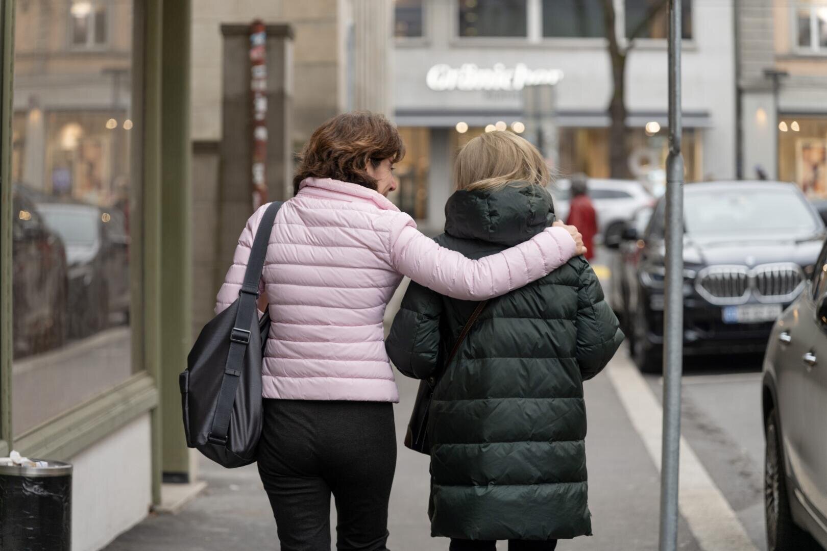 Zwei Menschen gehen einen Bürgersteig entlang, einer hat den Arm um die Schulter des anderen gelegt. Beide tragen Pufferjacken, eine rosa und eine dunkelgrüne, und haben Taschen dabei. Autos und Schaufenster säumen die Straße.