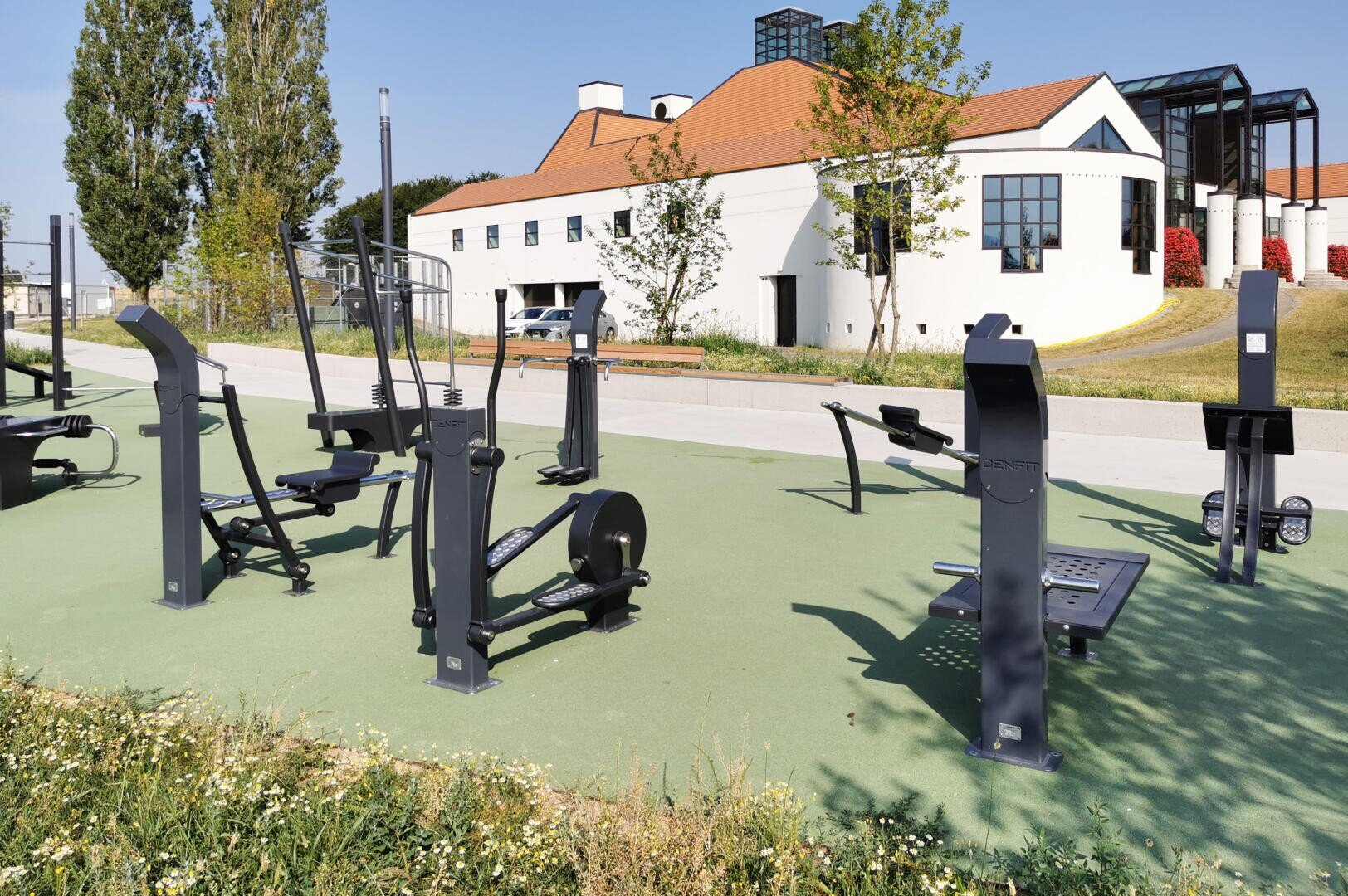 Fitnessstudio im Freien mit verschiedenen schwarzen Fitnessgeräten auf einer grünen Fläche, in der Nähe eines modernen weißen Gebäudes mit rotem Dach und großen Fenstern; Bäume und Gras umgeben das Gelände unter einem klaren Himmel.