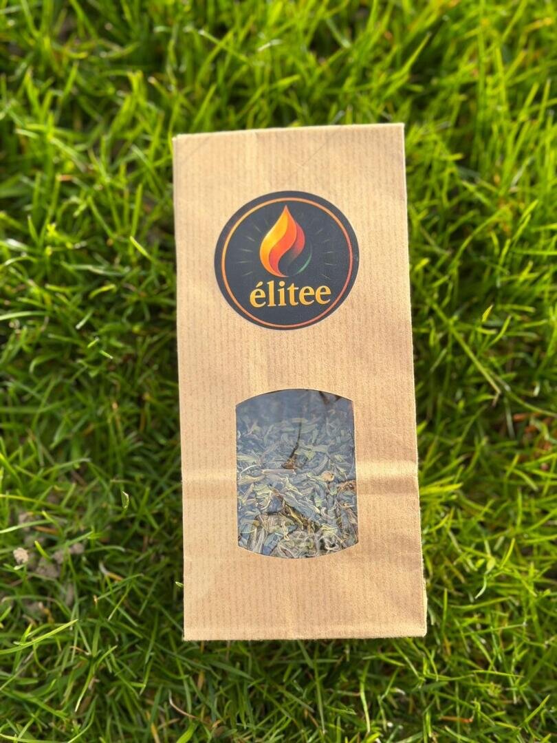 Ein braunes Papierpaket mit der Aufschrift éliteé und einem Flammenlogo liegt auf grünem Gras. Die Verpackung hat ein durchsichtiges Fenster, in dem getrocknete Blätter oder Kräuter zu sehen sind.