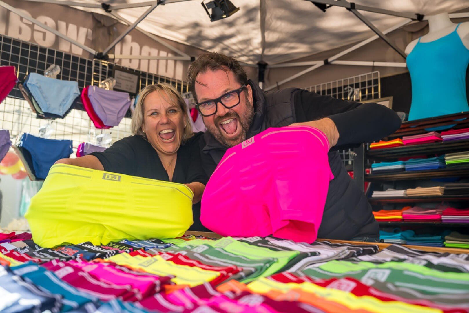 Zwei fröhliche Menschen halten an einem Marktstand knallbunte Unterwäsche in die Höhe, eine neongelbe und eine pinke, während sie hinter einer Auslage mit verschiedenfarbiger Unterwäsche auf einem Tisch stehen.