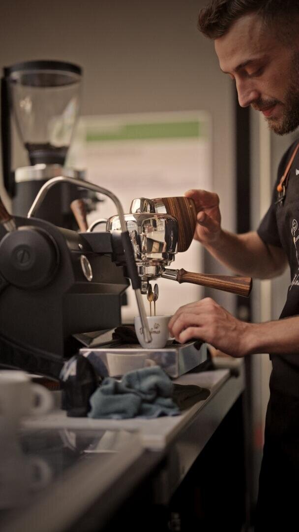 Ein Barista bedient eine Espressomaschine und gießt den Kaffee in eine Tasse, während auf dem Tresen eine Kaffeemühle und andere Kaffeegeräte zu sehen sind.