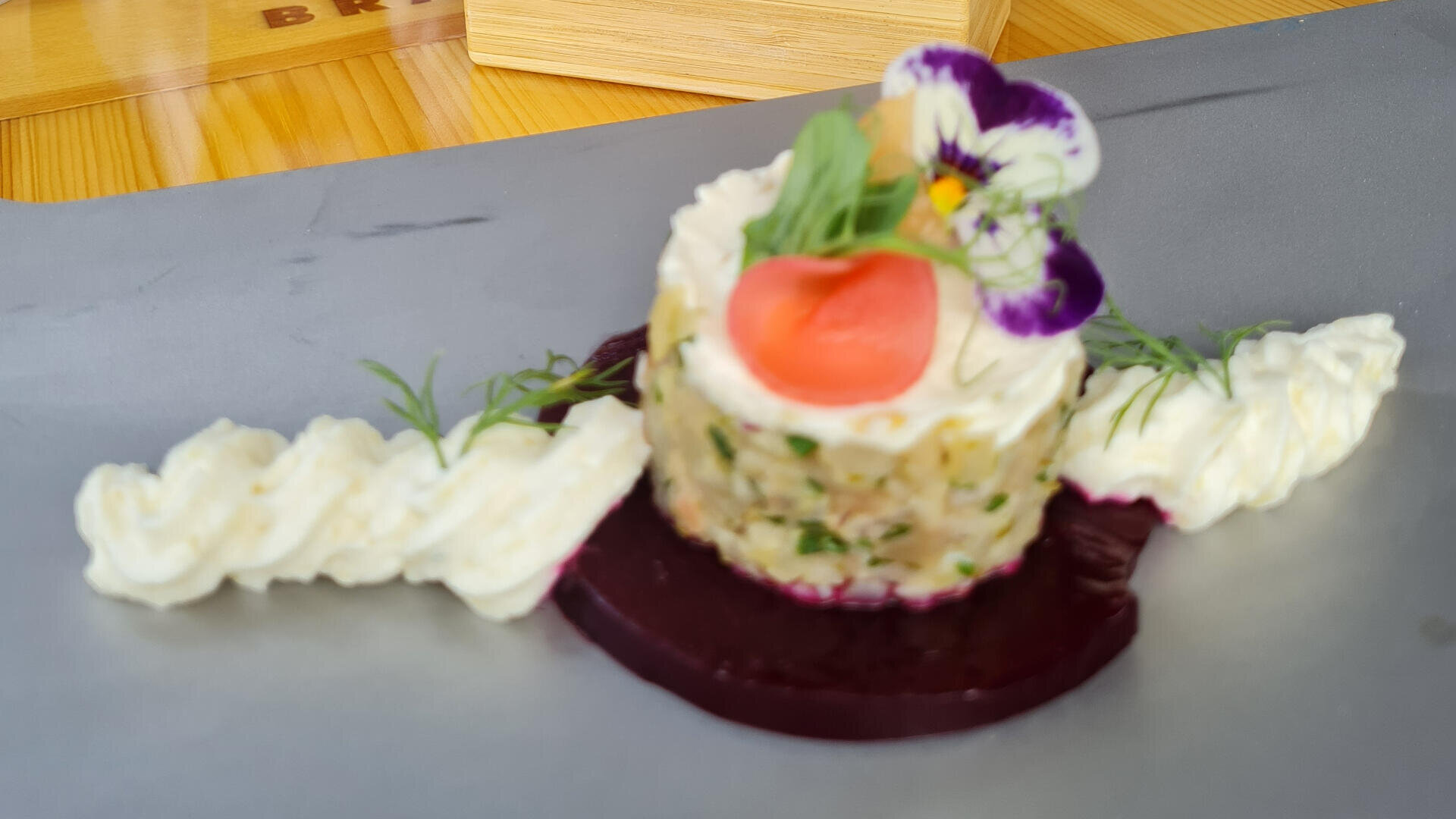 Ein Gourmetgericht mit einem runden Tartar, das mit einer Kirschtomate und einer essbaren Blume belegt ist, mit Kräutern garniert und auf einer Rübenscheibe serviert wird, mit Sahne oder Mousse an der Seite, präsentiert auf einem grauen Teller.