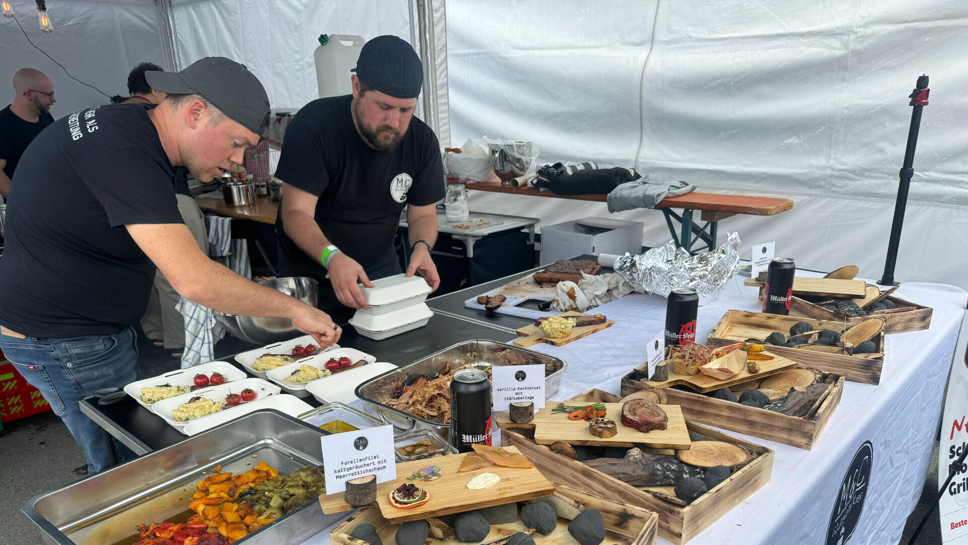 Zwei Männer in schwarzen Hemden bereiten hinter einem Tisch mit verschiedenen Fleisch-, Käse- und Gemüsesorten an einem Essensstand in einem weißen Zelt Boxen zum Mitnehmen vor. Auf dem Tisch stehen auch Schilder und Getränkedosen.