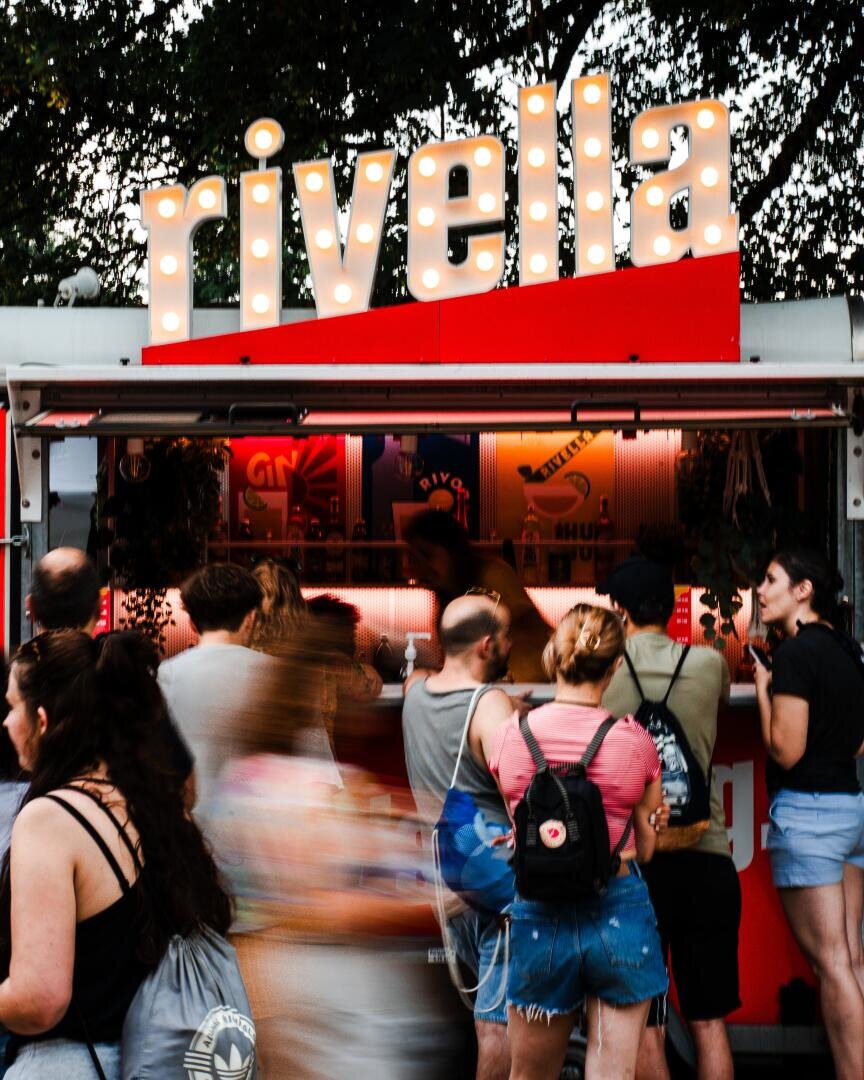 Eine Gruppe von Menschen steht vor einem Imbisswagen mit einem großen, beleuchteten Nietenschild an der Spitze. Im Hintergrund sind Bäume zu sehen, was auf eine Veranstaltung oder ein Fest im Freien hindeutet.
