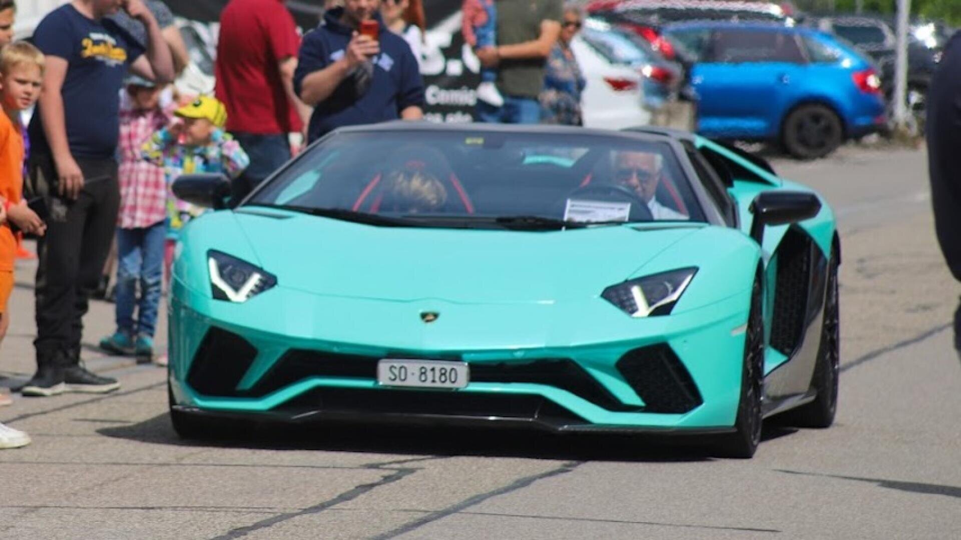 Ein türkisfarbenes Lamborghini-Cabriolet fährt eine Straße entlang, die von Menschen gesäumt ist, von denen einige Fotos machen und andere zusehen. Die Umgebung besteht aus geparkten Autos und einer hellen, sonnigen Atmosphäre.