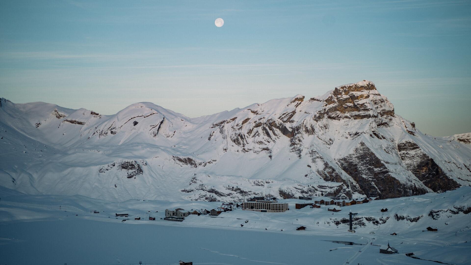 Schneebedeckte Berge bei klarem Himmel, über denen der Mond zu sehen ist. Am Fuße der Berge befinden sich Gebäude, im Vordergrund ein gefrorener See oder eine verschneite Ebene.