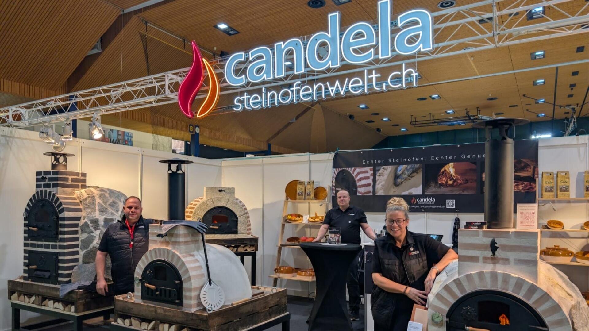 Drei Personen stehen an einem Messestand von candela steinofenwelt.ch und stellen verschiedene Steinöfen aus. Der Stand hat eine grosse Leuchtreklame, ausgestellte Öfen und eine Holzdecke darüber.