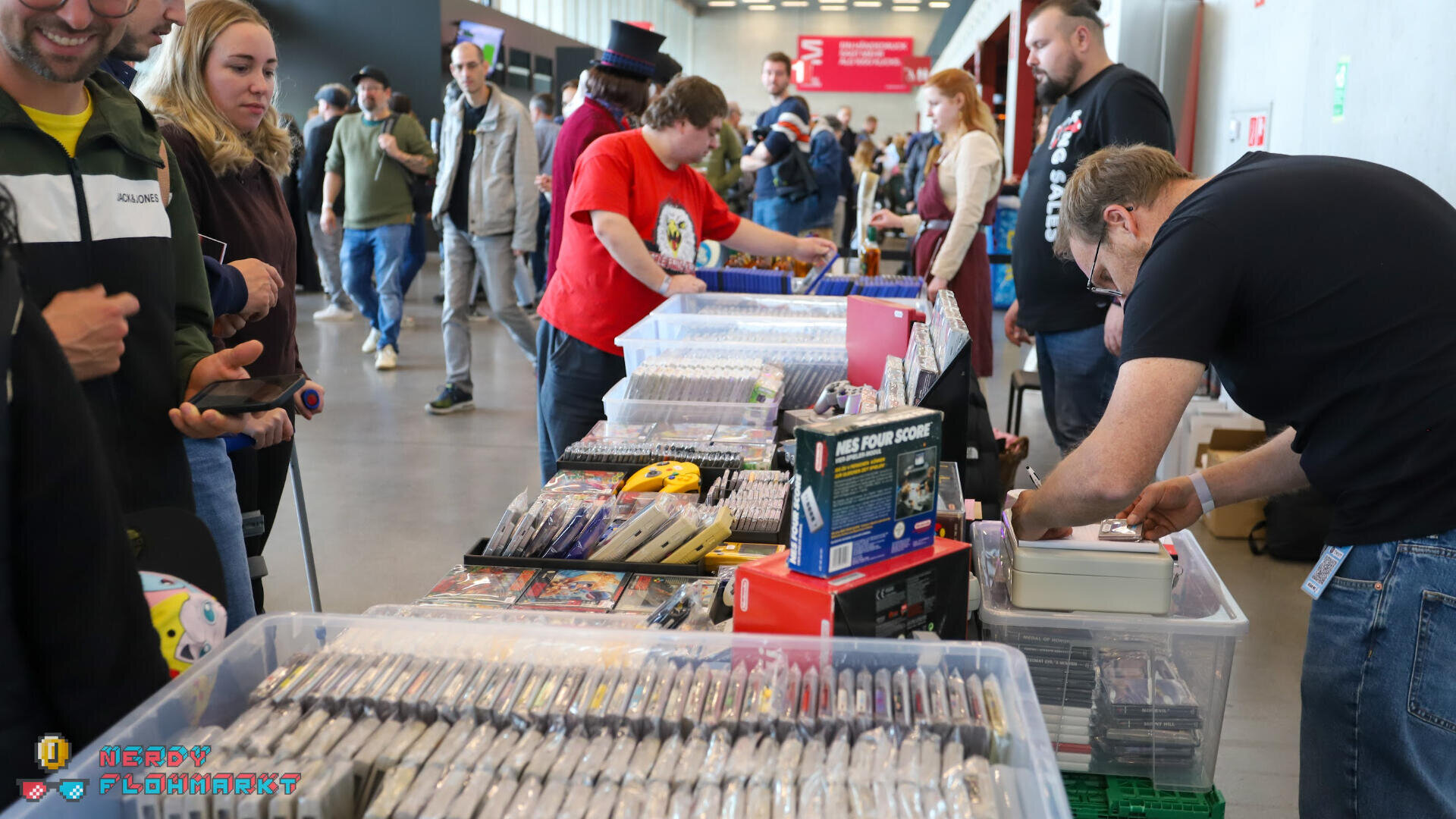 Die Leute stöbern und kaufen Retro-Videospiele und Zubehör an einem Verkäufertisch in einer belebten Kongresshalle. Der Tisch ist mit Kassettenhüllen, Videospielen und Controllern gefüllt. Käufer und Verkäufer tauschen sich in einer lebhaften Atmosphäre aus.